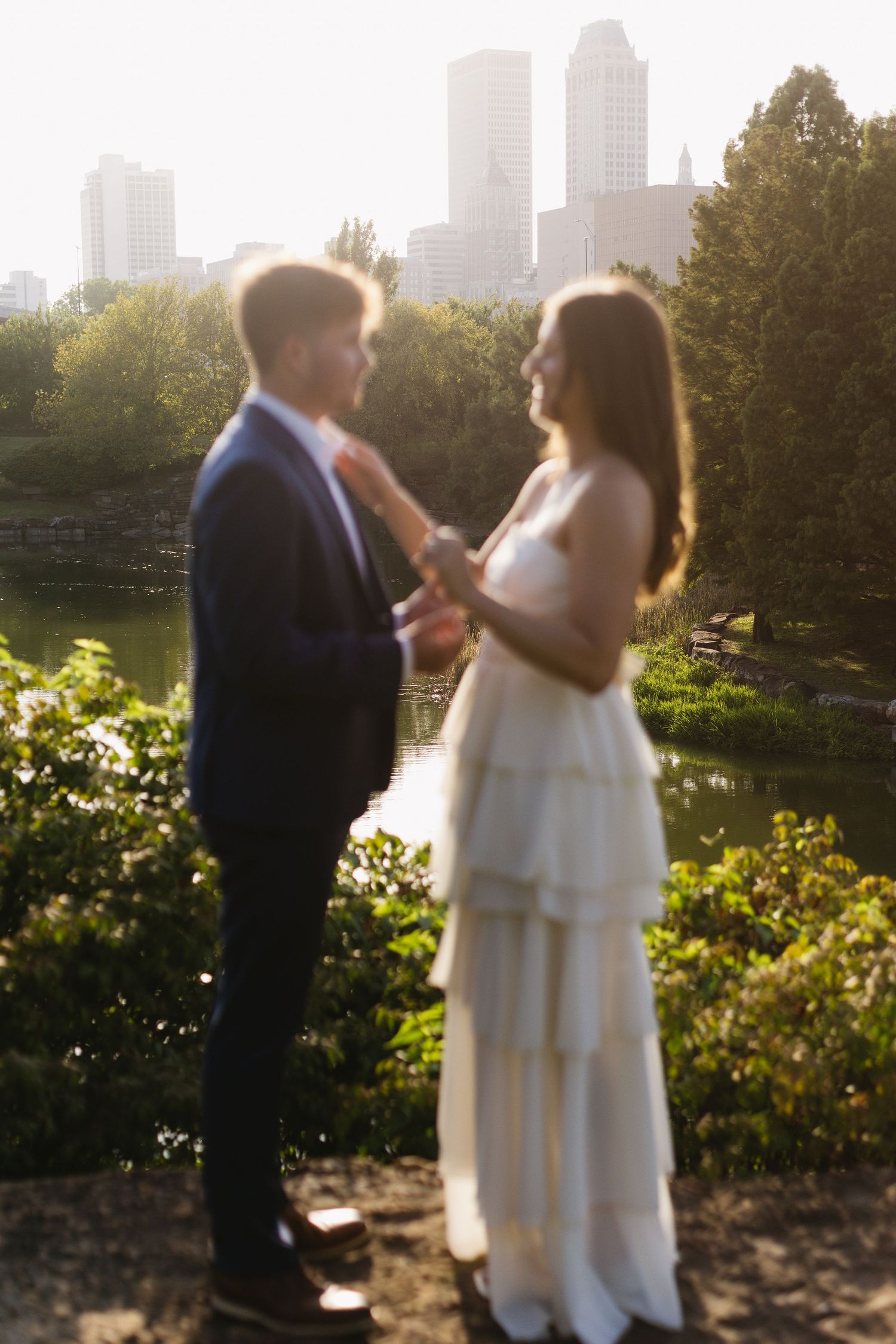 A couple shares an intimate moment in a garden during golden hour with city buildings visible in the background.
