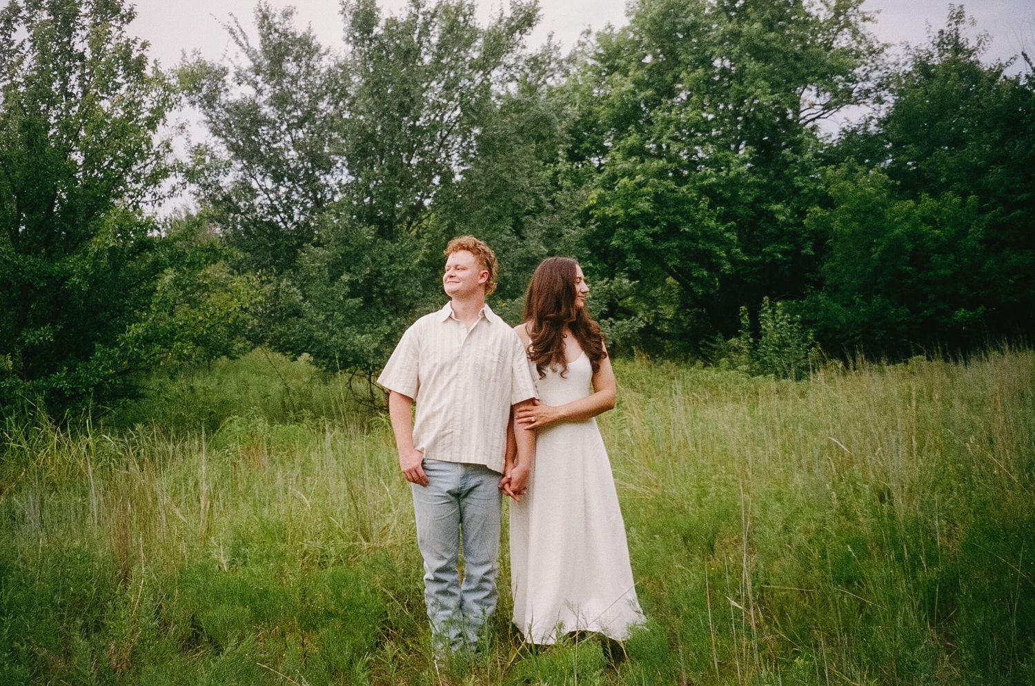 A couple in romantic attire walks together through a lush summer meadow on an overcast day.