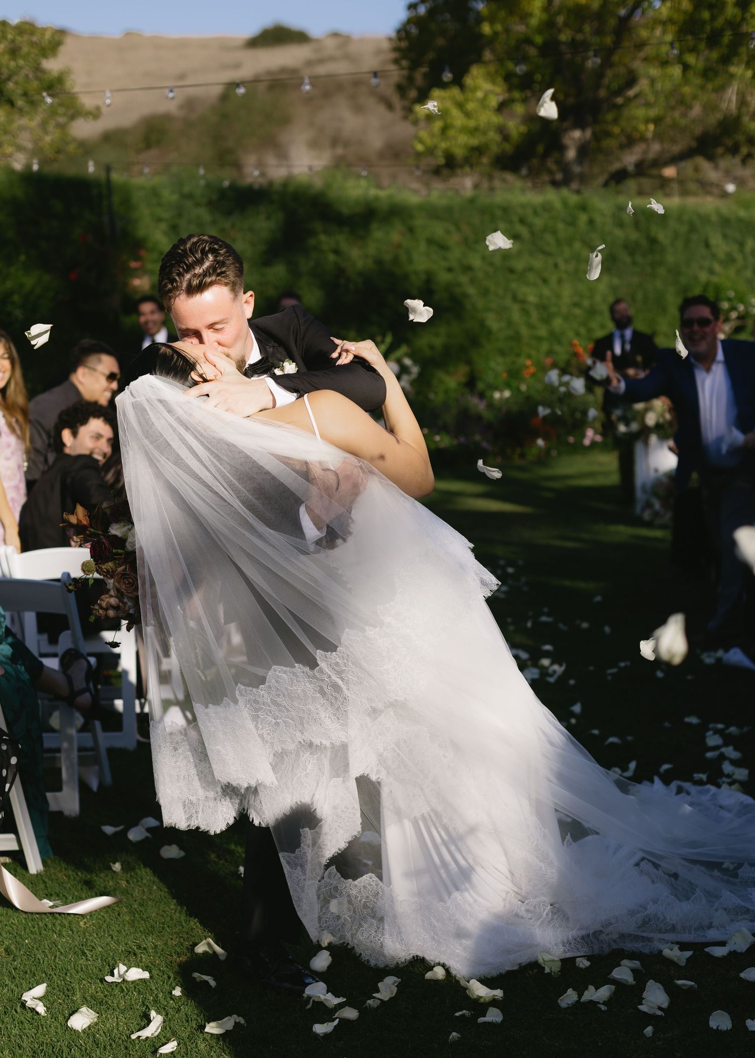 A bride and groom share a romantic dip dance move as white flower petals fall around them during an outdoor wedding celebration.