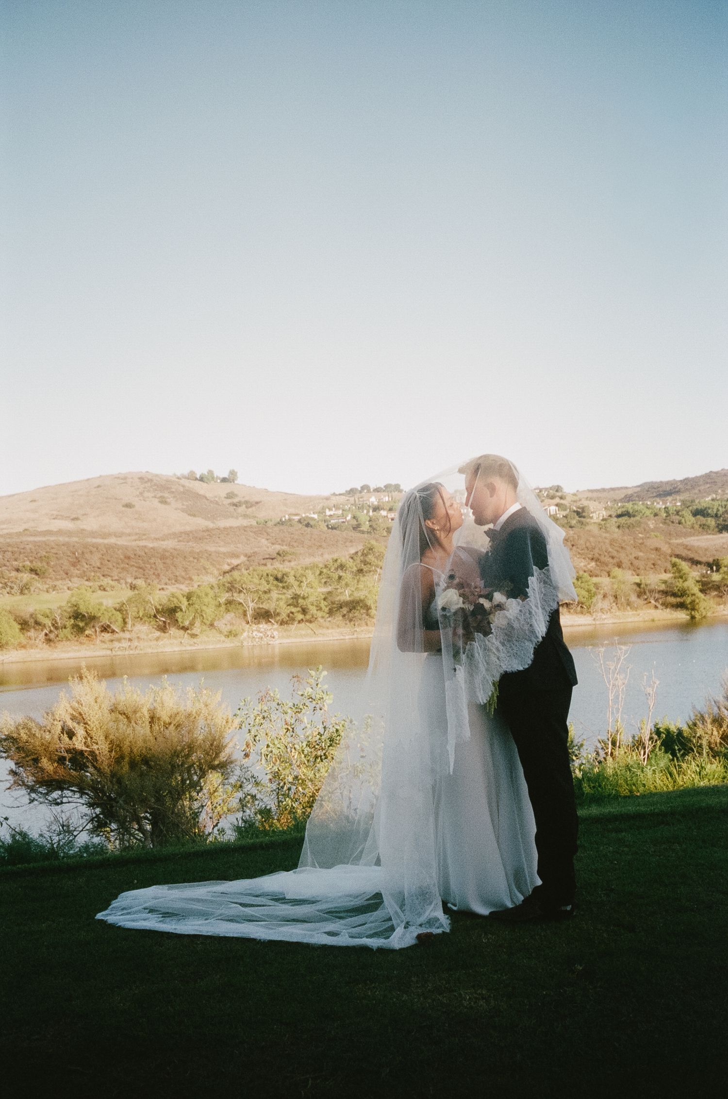 A romantic wedding couple shares a moment at sunset by a serene lake with mountains in the background.