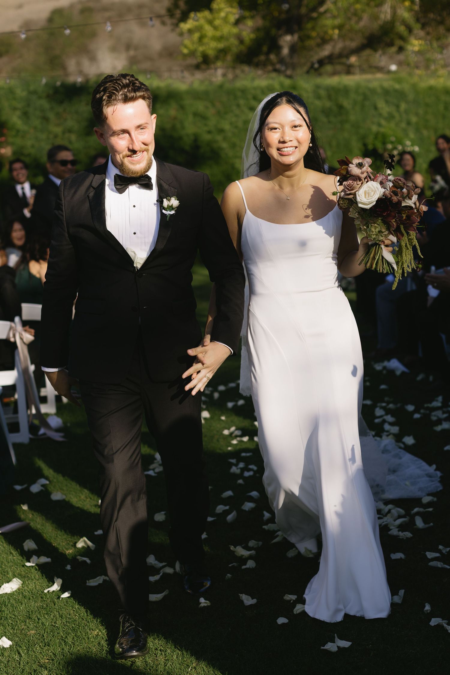 A newlywed couple walks down a petal-strewn aisle after their outdoor wedding ceremony.