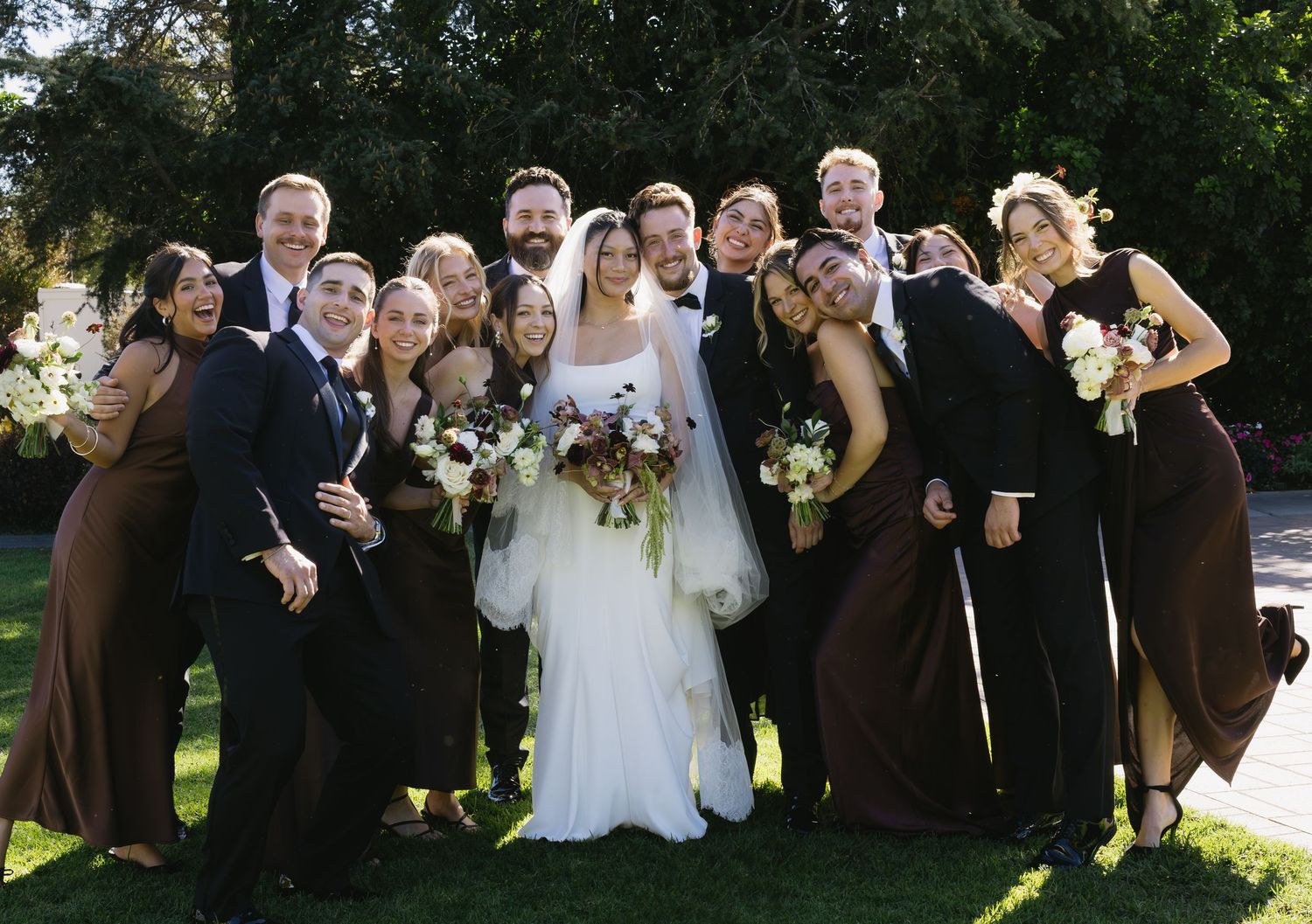 Wedding party group photo outdoors with guests in dark attire.