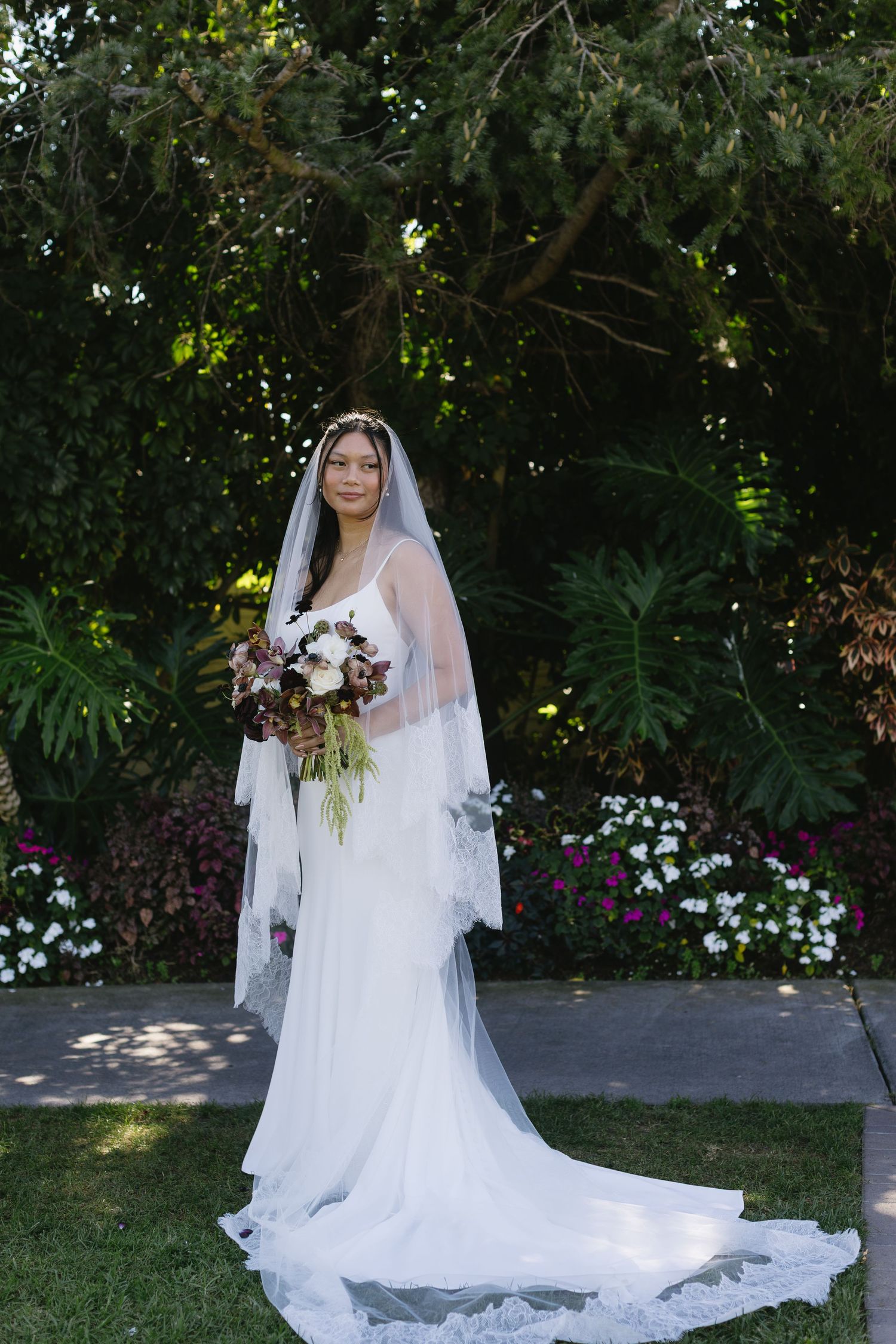 Bride in flowing white gown and veil poses in garden setting with lush greenery and flowers.