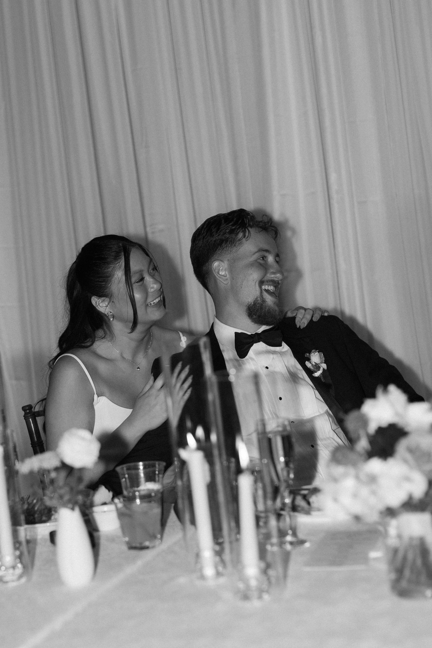 Black and white candid reception photo showing guests laughing at dinner table.