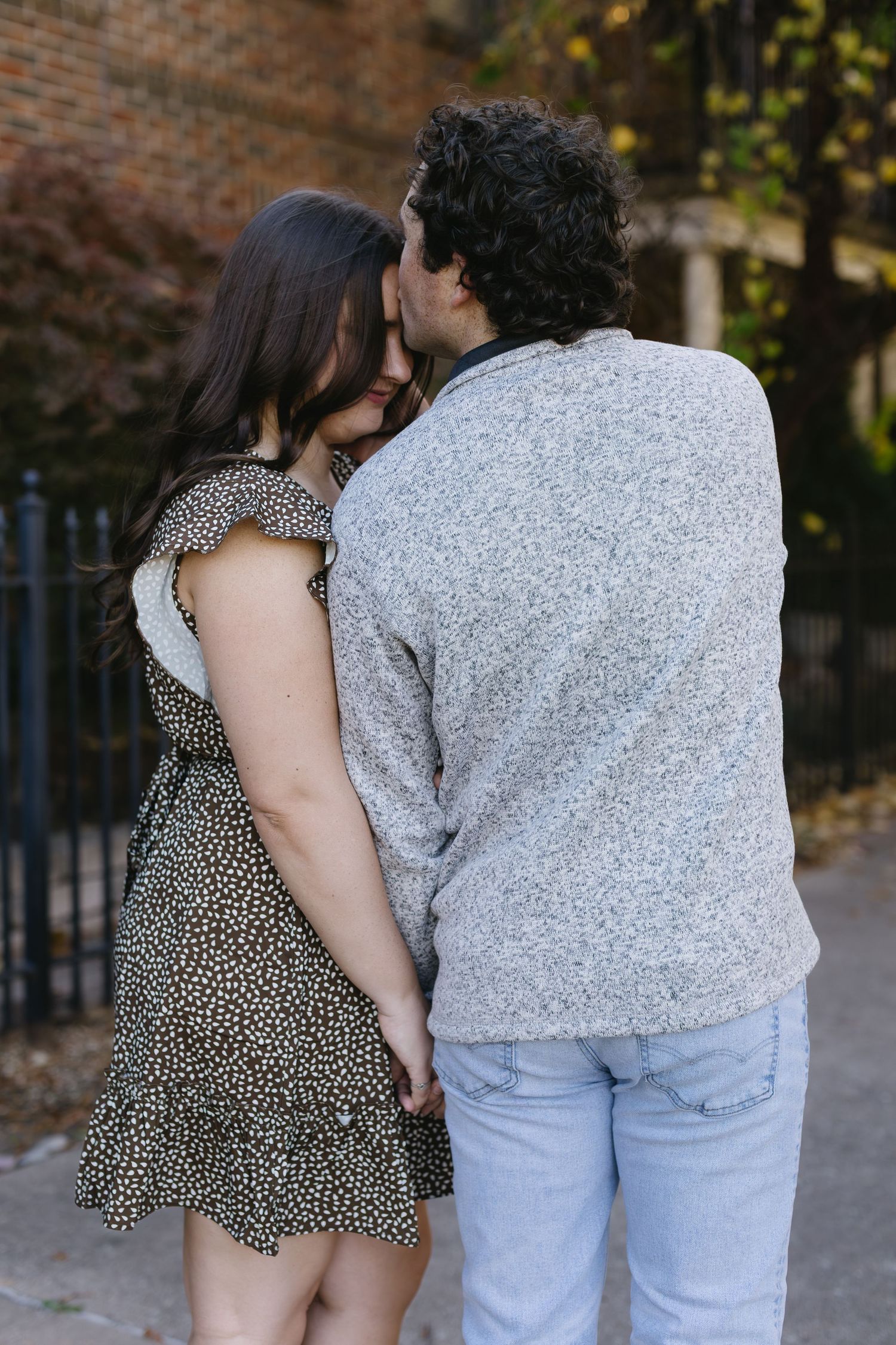 A couple in casual attire embraces while posing against a brick wall backdrop.