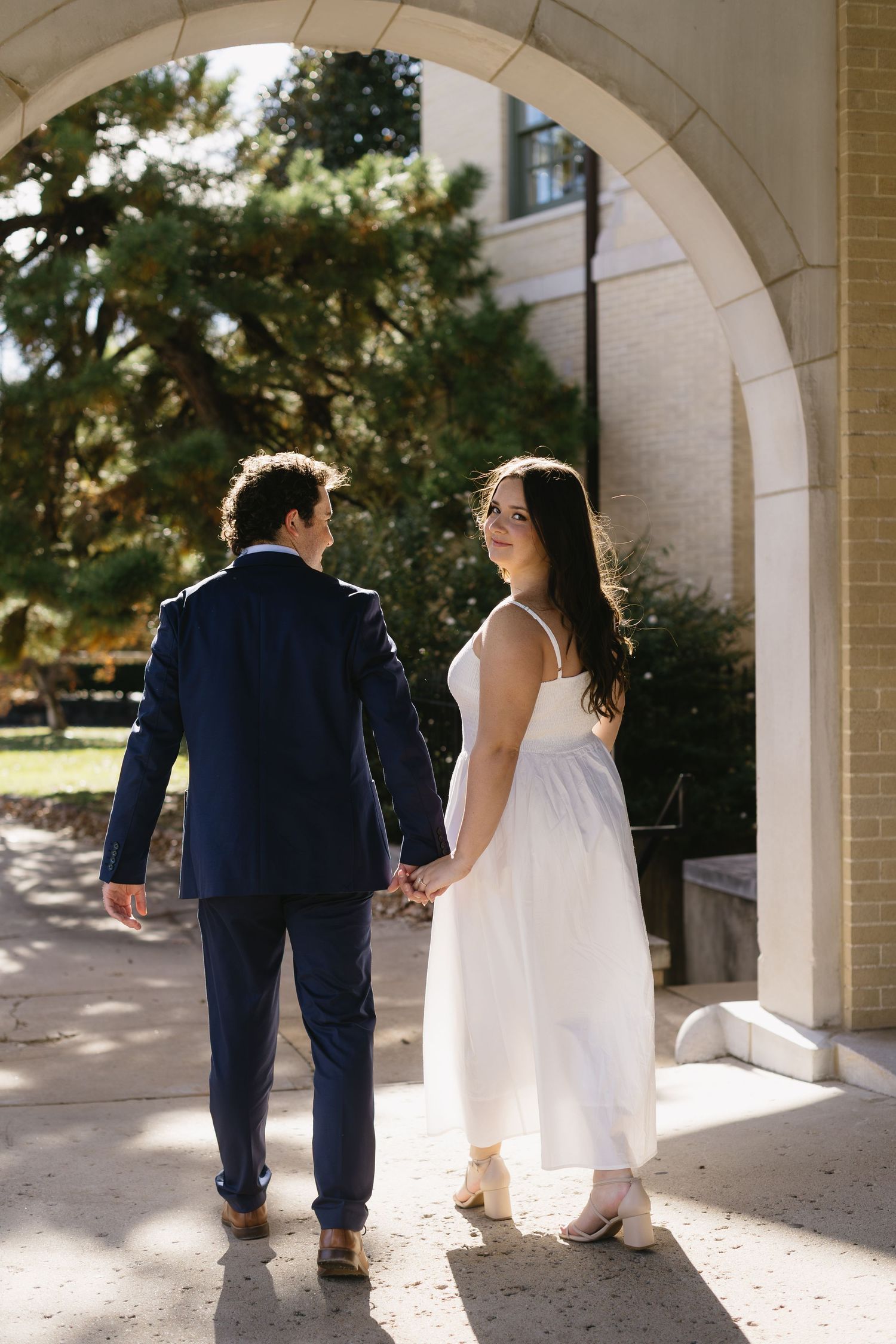 A couple in wedding attire sits together under a stone archway in warm sunlight.