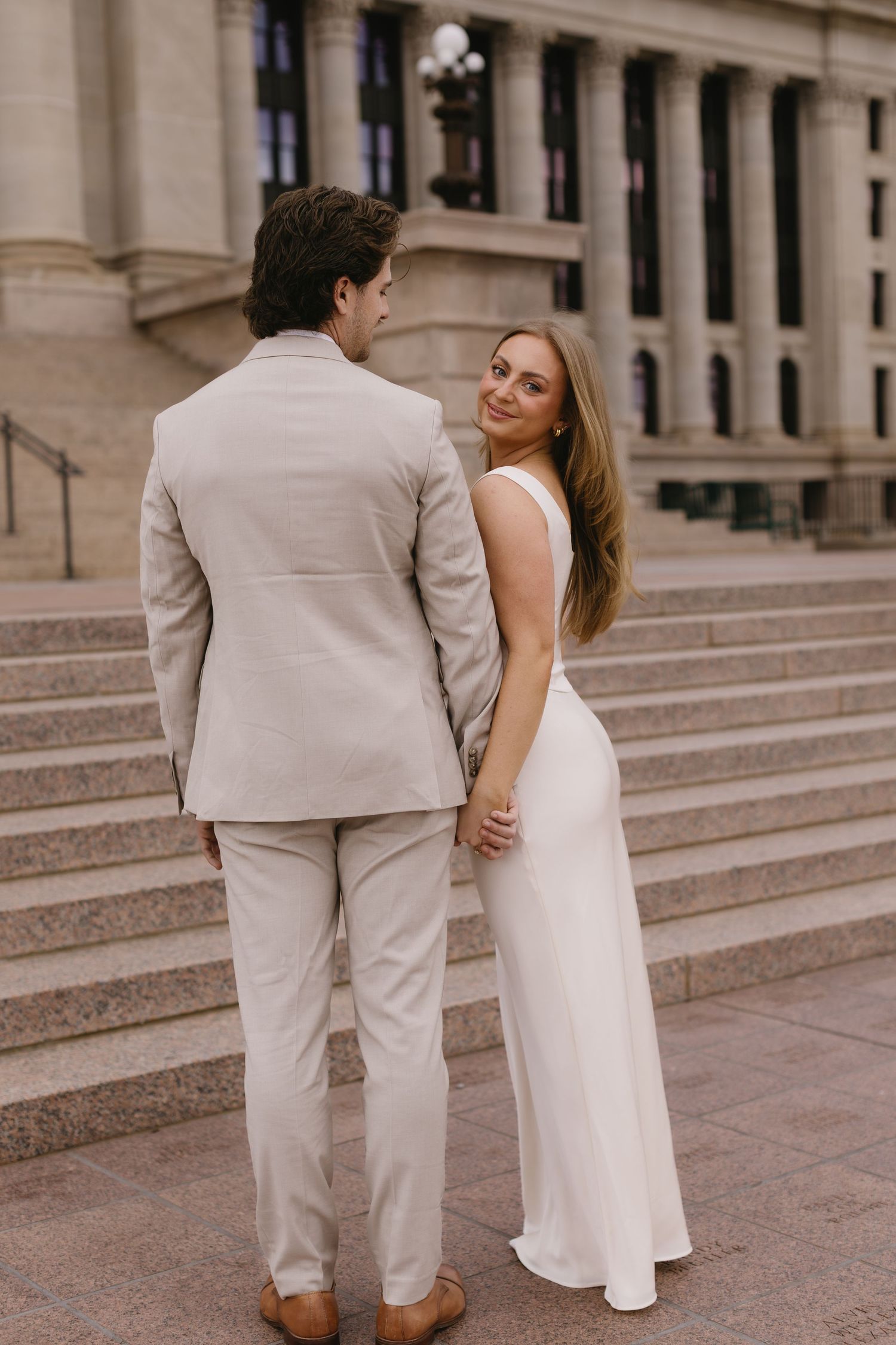 A couple in light colored formal wear ascends stone steps together at a stately venue.