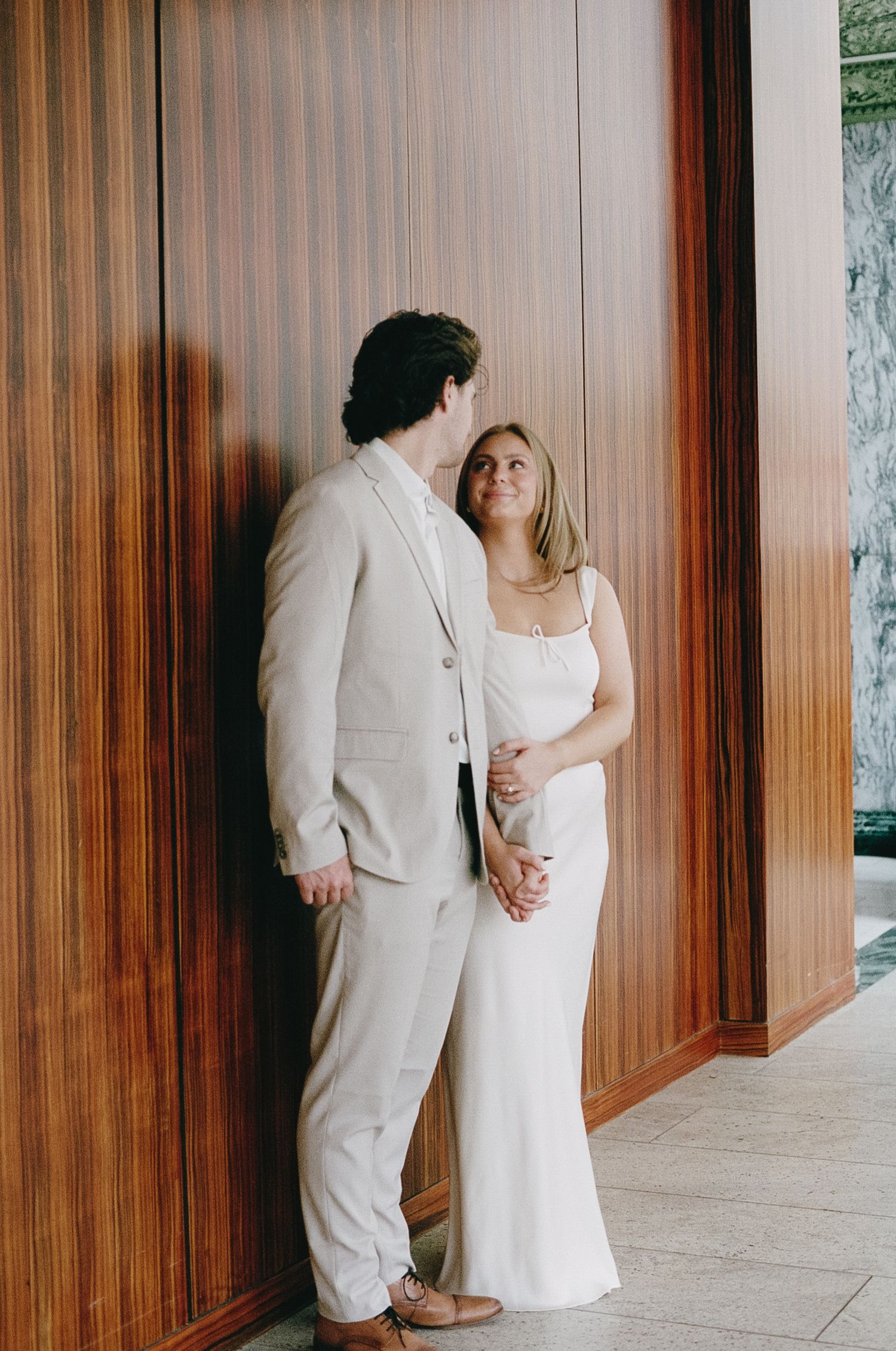 A couple in matching formal attire stands together against a wooden paneled wall in an elegant setting.