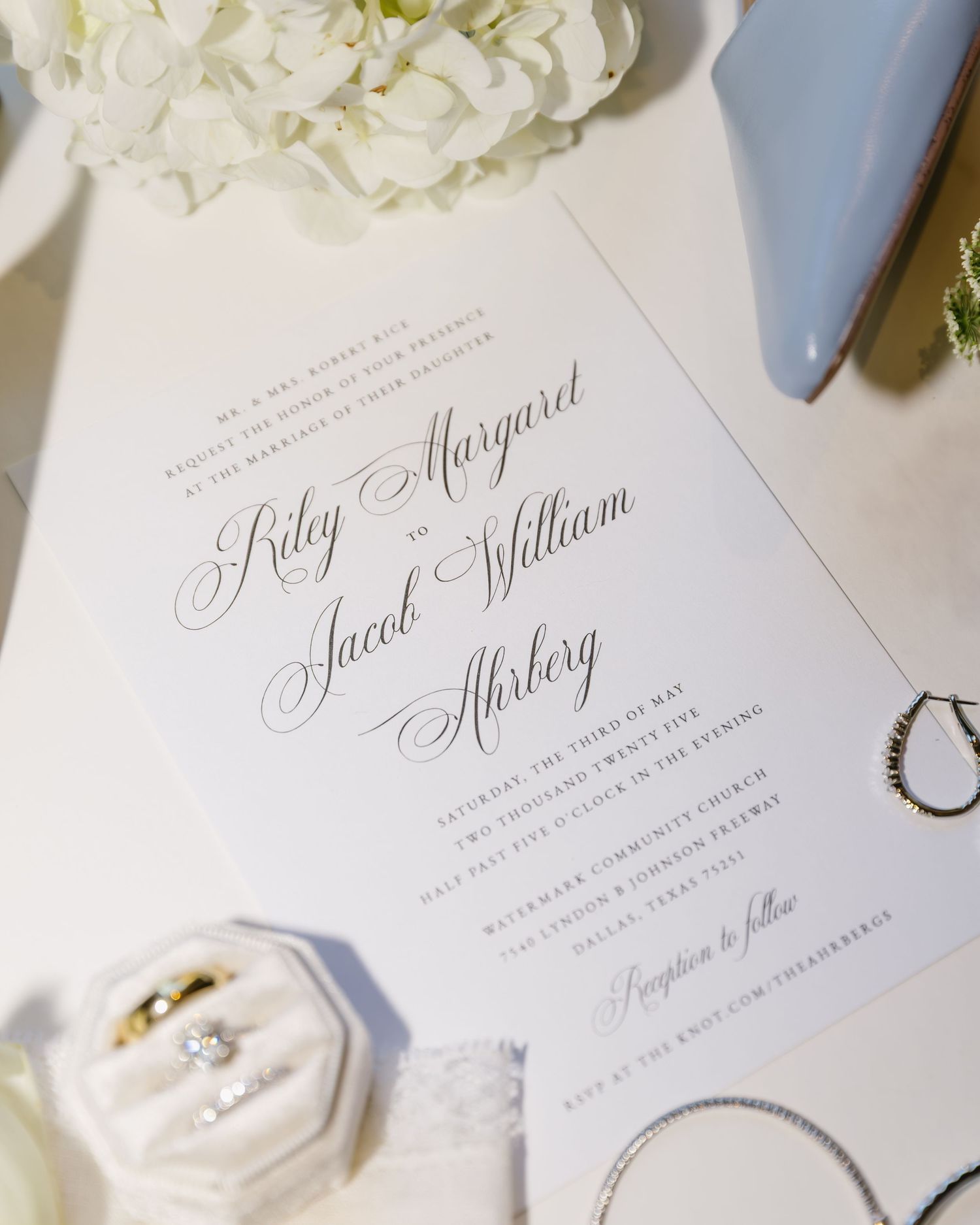 Elegant white wedding invitation suite with calligraphy script text, ring, and floral accents on light background.