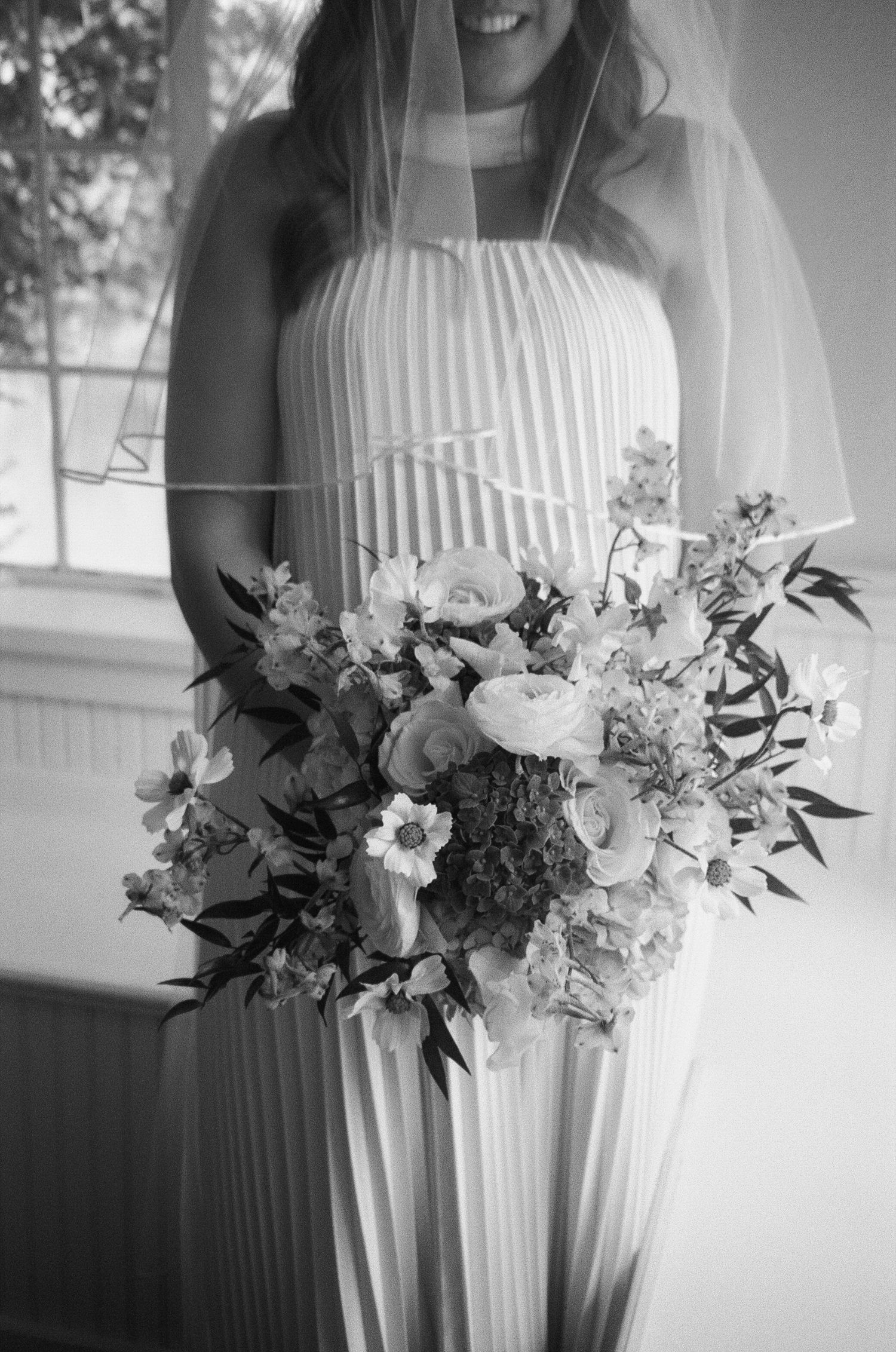 Beautiful wedding bouquet of mixed white flowers and greenery held against an elegant pleated white dress in black and white.