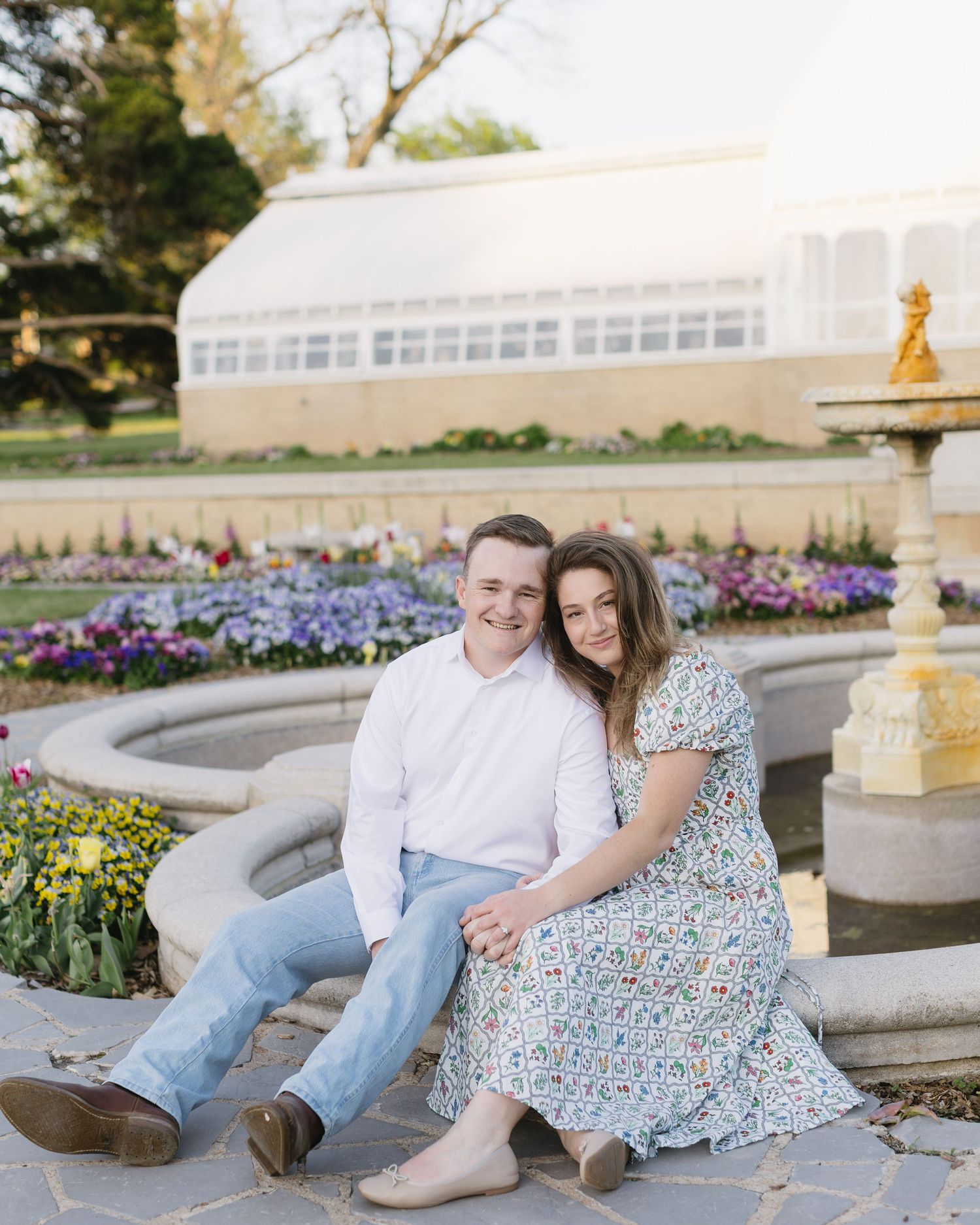 A couple relaxes by an ornate fountain surrounded by purple flowers in a garden setting.
