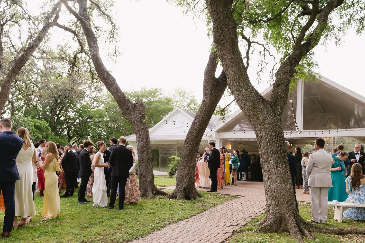 Wedding guests gather outdoors under large oak trees along a brick pathway at an elegant outdoor reception venue.