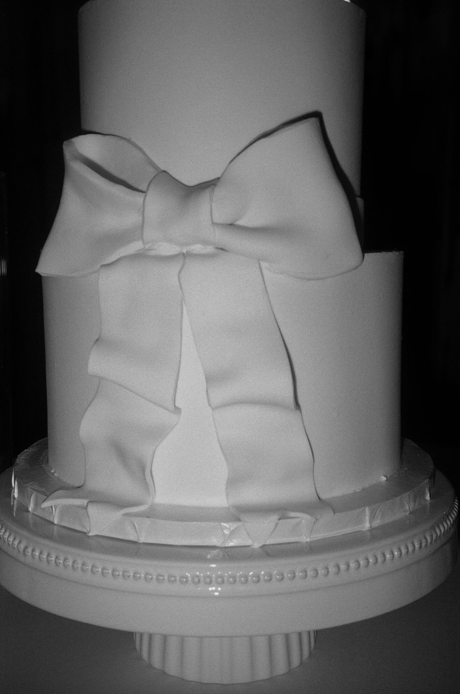 Elegant white fondant cake decorated with a large bow detail on white pedestal cake stand.