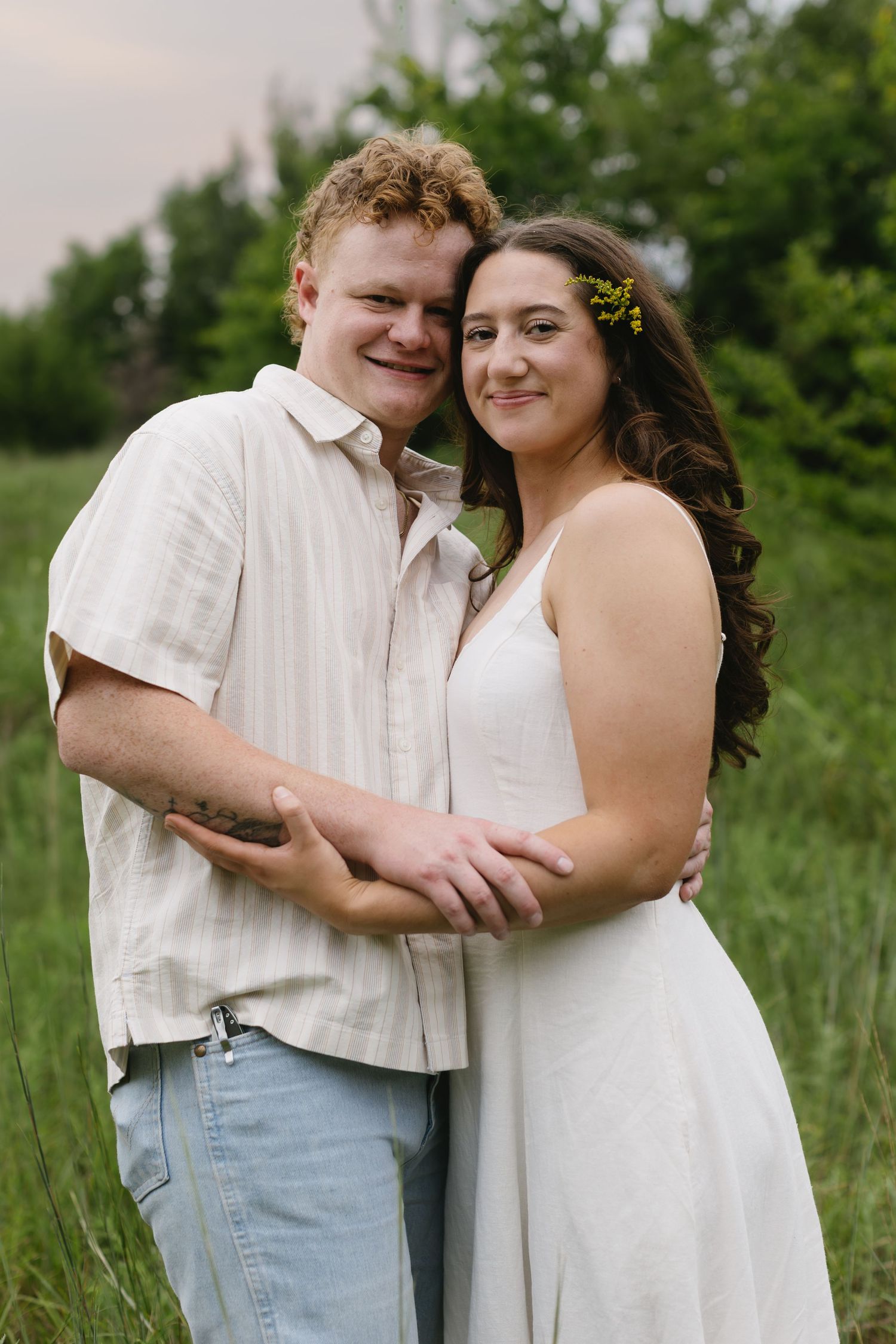 A couple in coordinating white outfits embrace lovingly in a summer field.