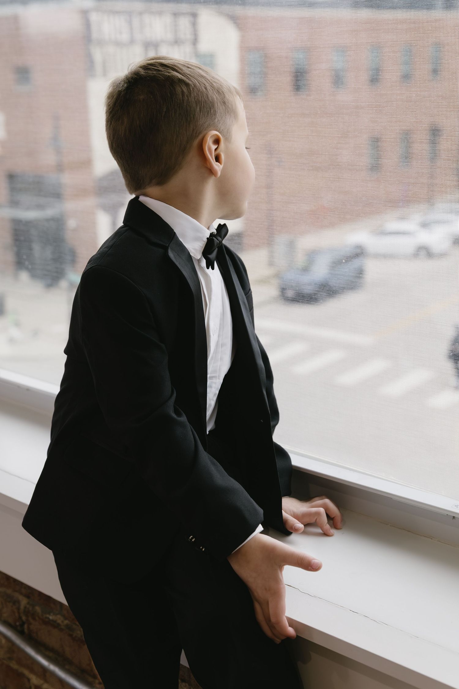 Young person in formal black suit gazing out window in contemplative pose.