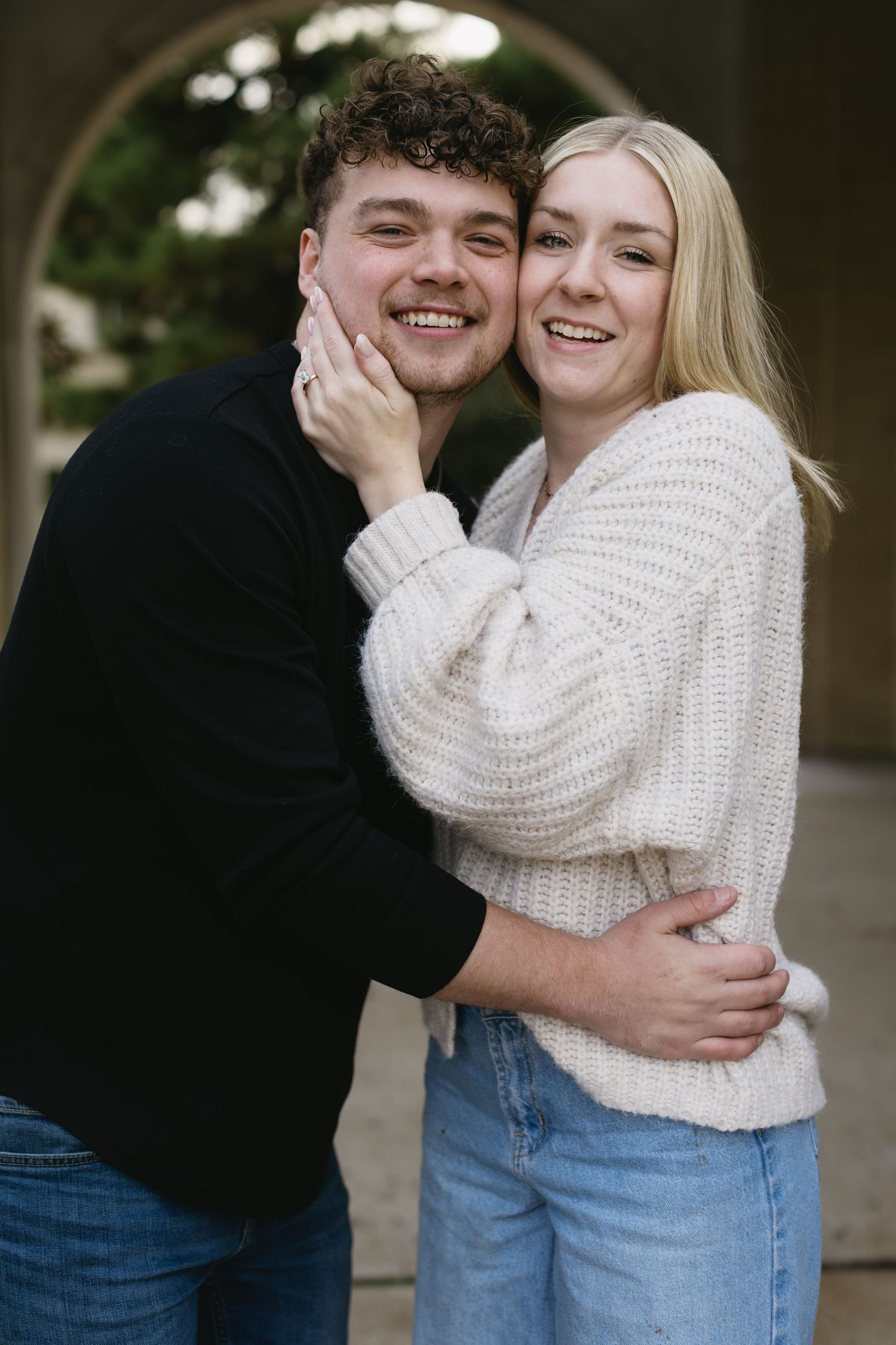 A couple embraces while wearing casual outfits of a black sweater and cream knit sweater with jeans outdoors.
