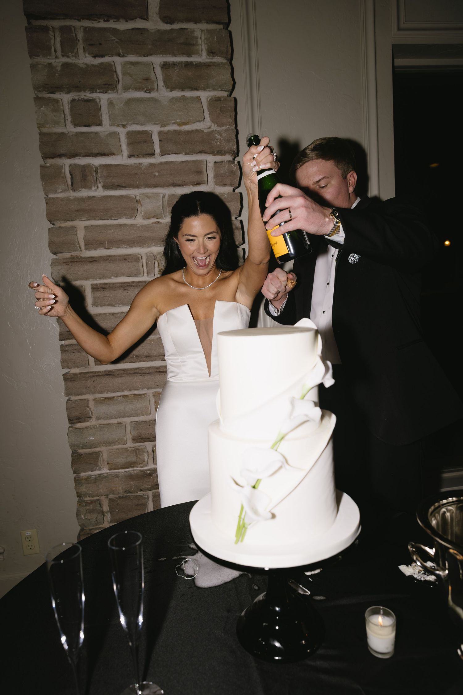 Newlyweds celebrating playfully next to their white tiered wedding cake.