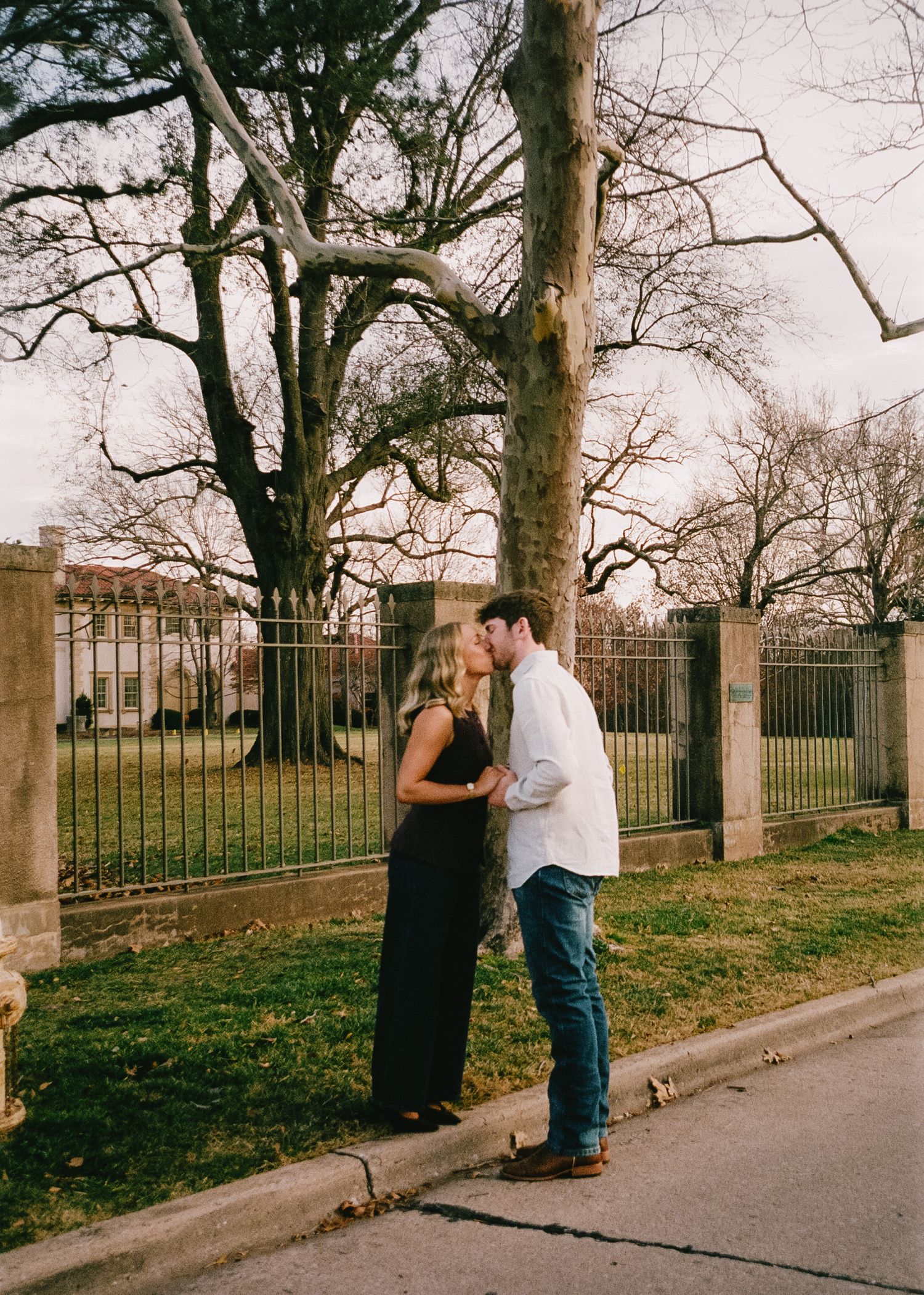 A couple sharing romantic moments along a tree-lined pathway with iron fencing.