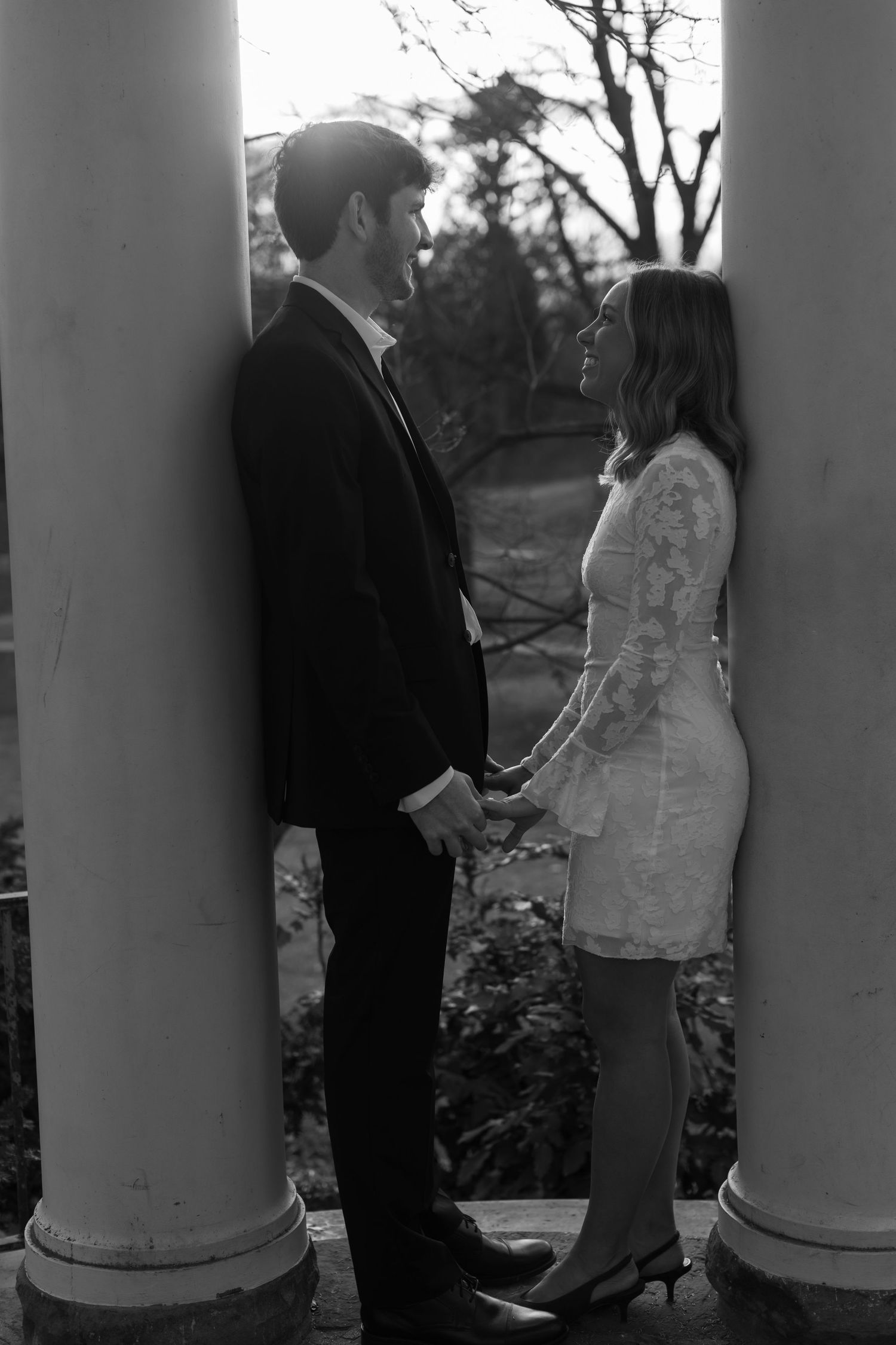A romantic couple poses between classical white columns in dramatic black and white lighting.