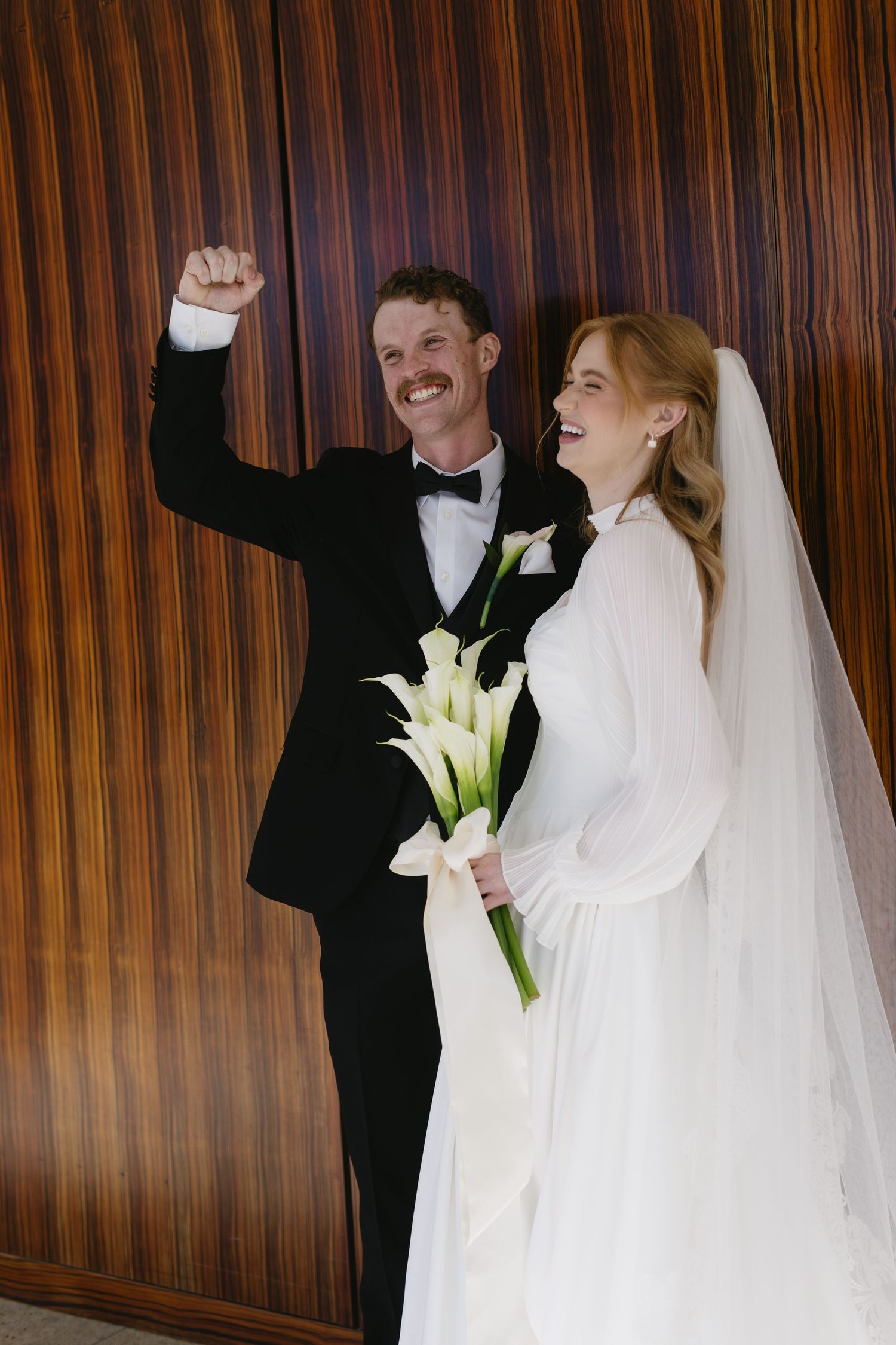 Wedding couple with calla lily bouquet celebrating joyfully against wood paneled wall.
