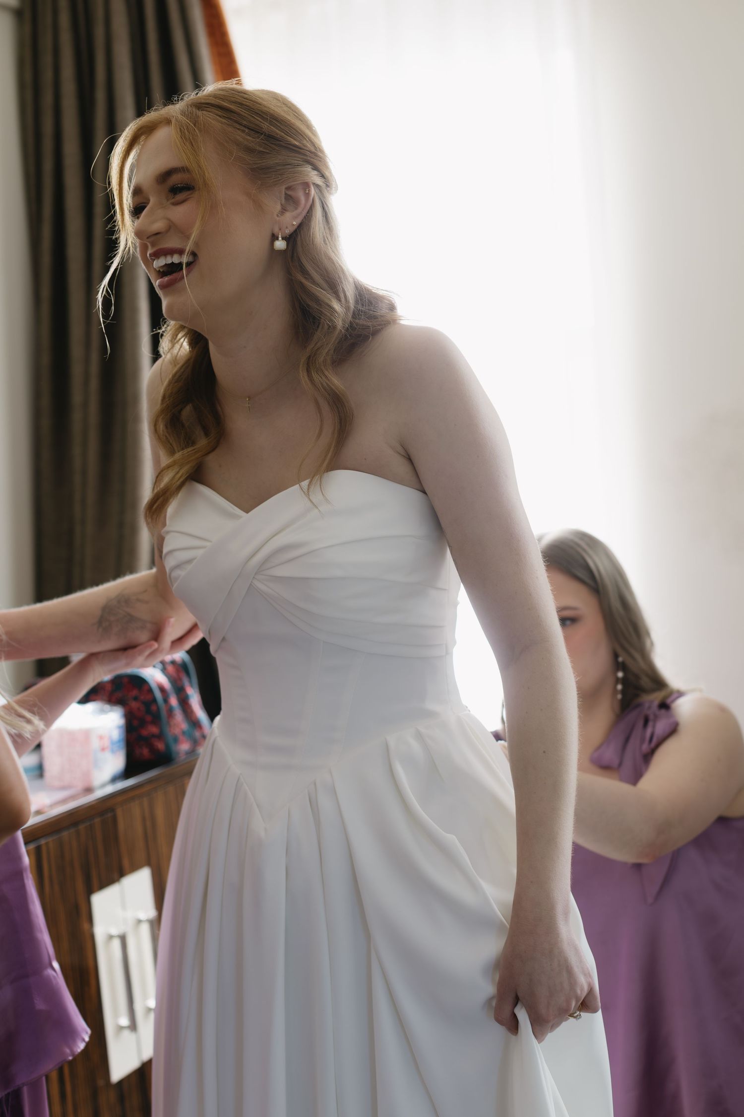 A bride in a flowing white dress gets ready in a sunlit room with purple accents.