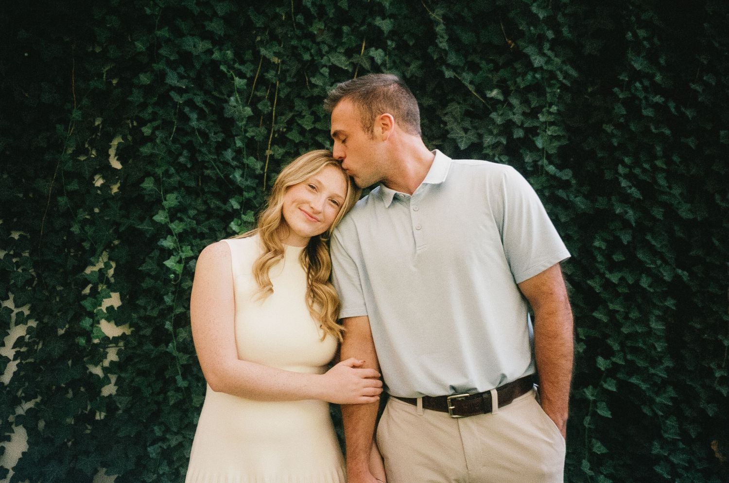 A couple shares a tender moment against a lush green ivy wall backdrop during an engagement photo session.