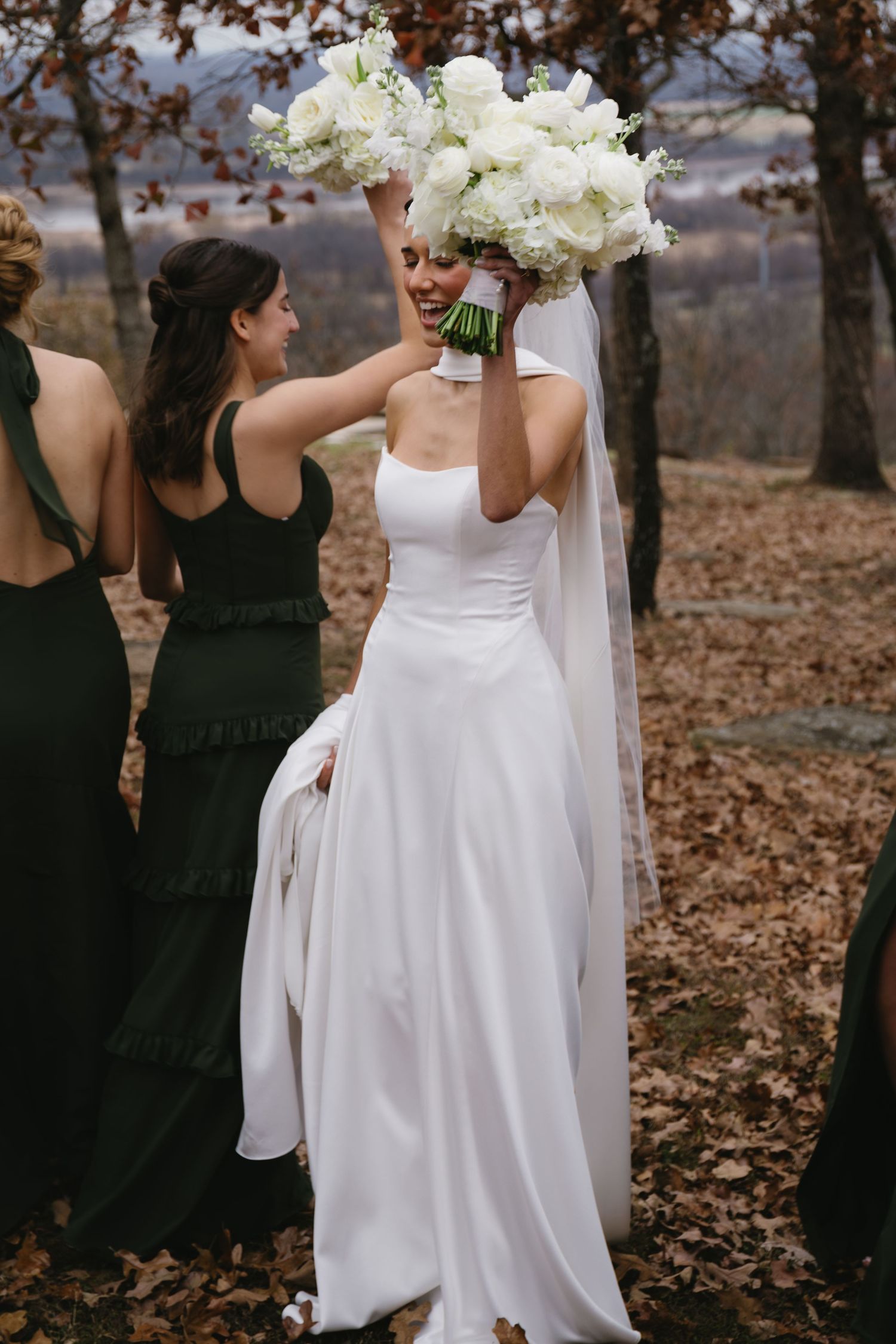 Bridesmaids in black dresses accompany a bride holding a white bouquet during an autumn outdoor wedding.
