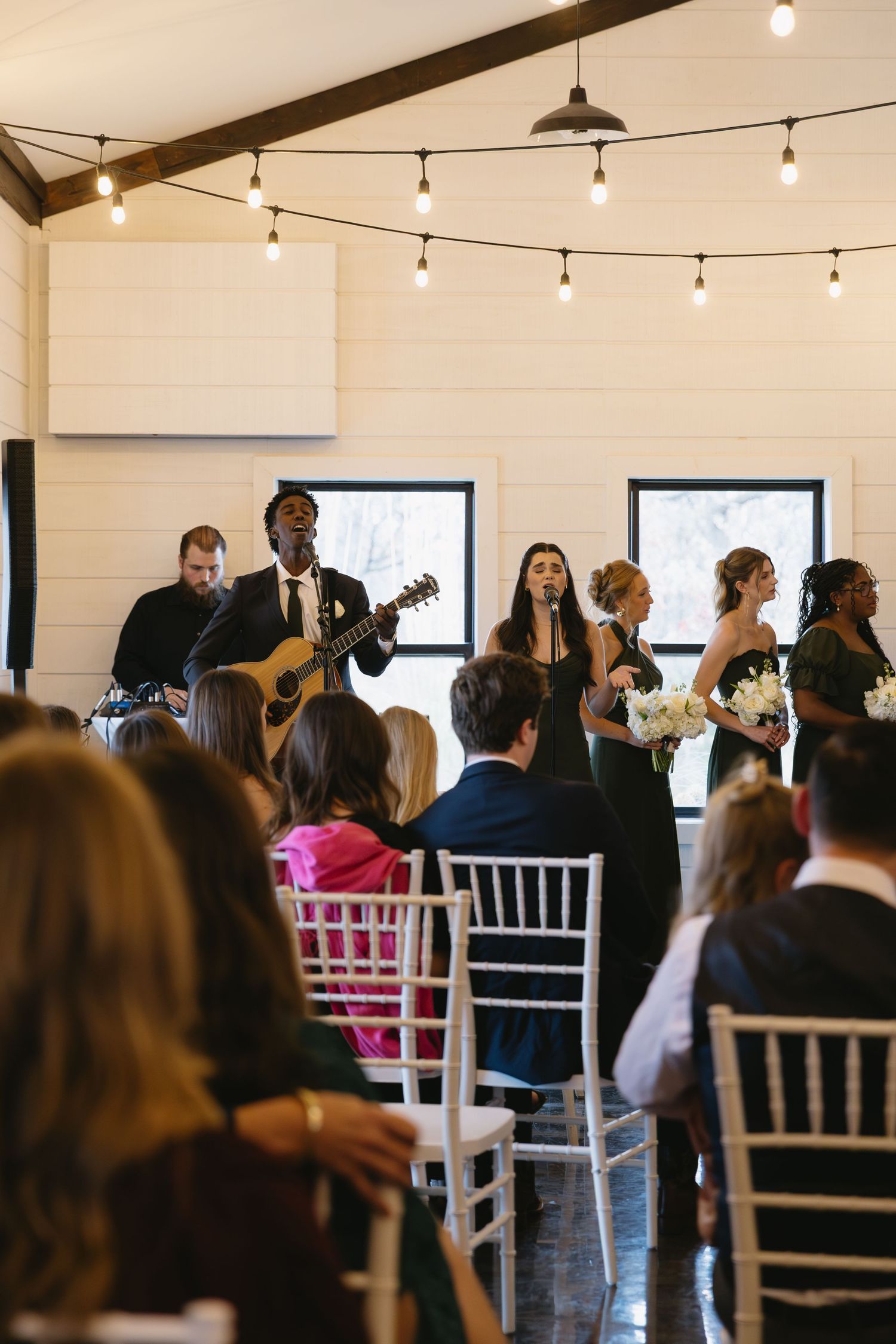 Live band performs at an indoor wedding ceremony with string lights and white shiplap walls.