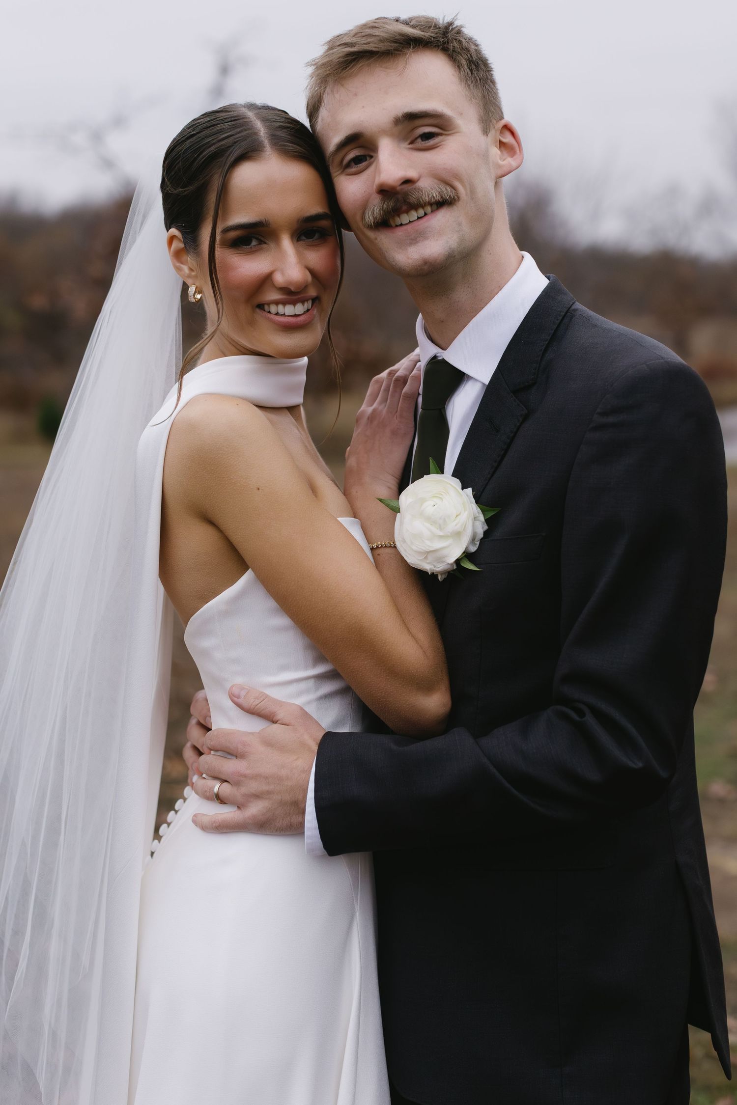 A wedding couple shares an intimate moment outdoors with the bride wearing a sleek white dress and veil.