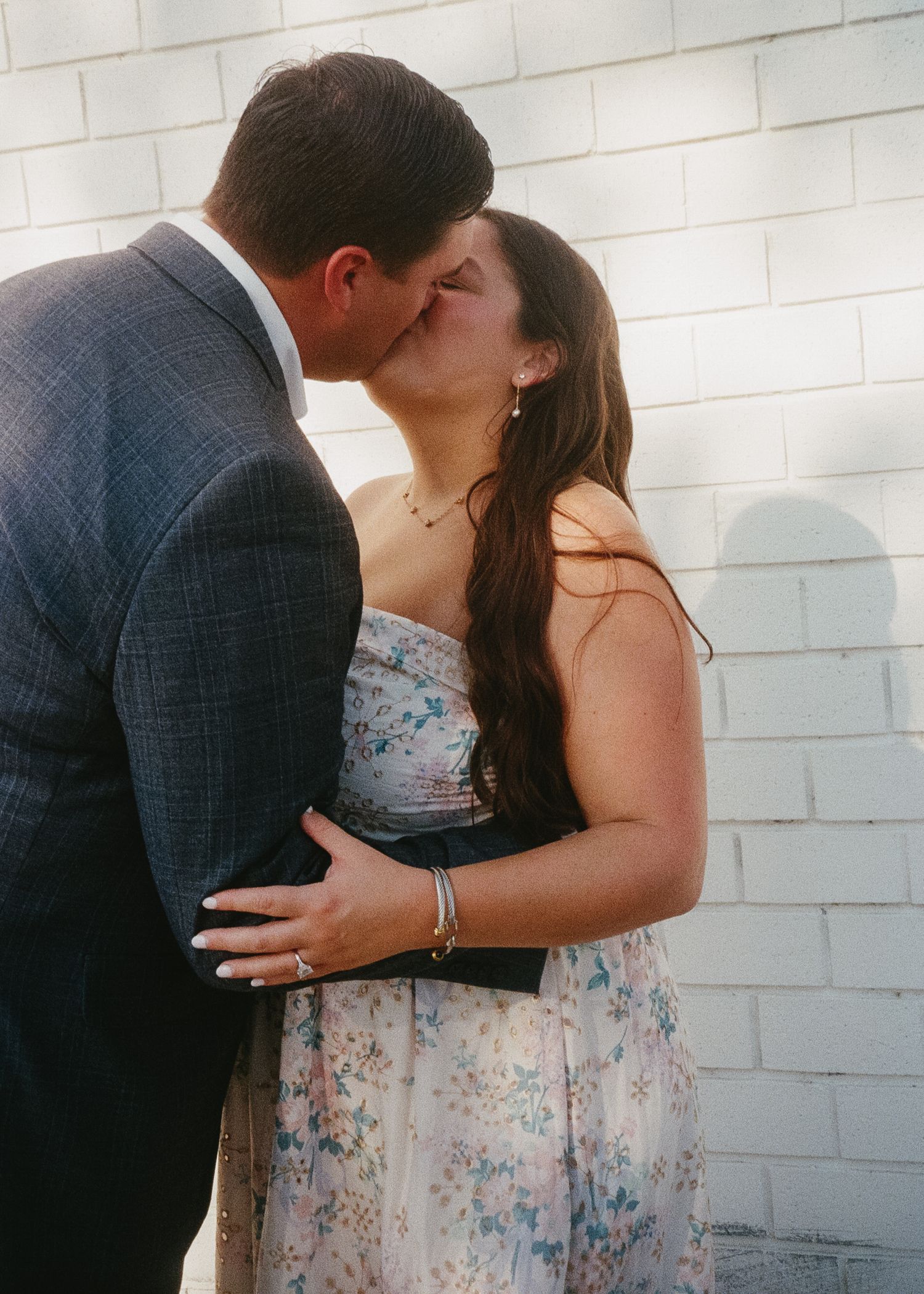 A couple in formal attire sharing a romantic moment against a white brick wall.