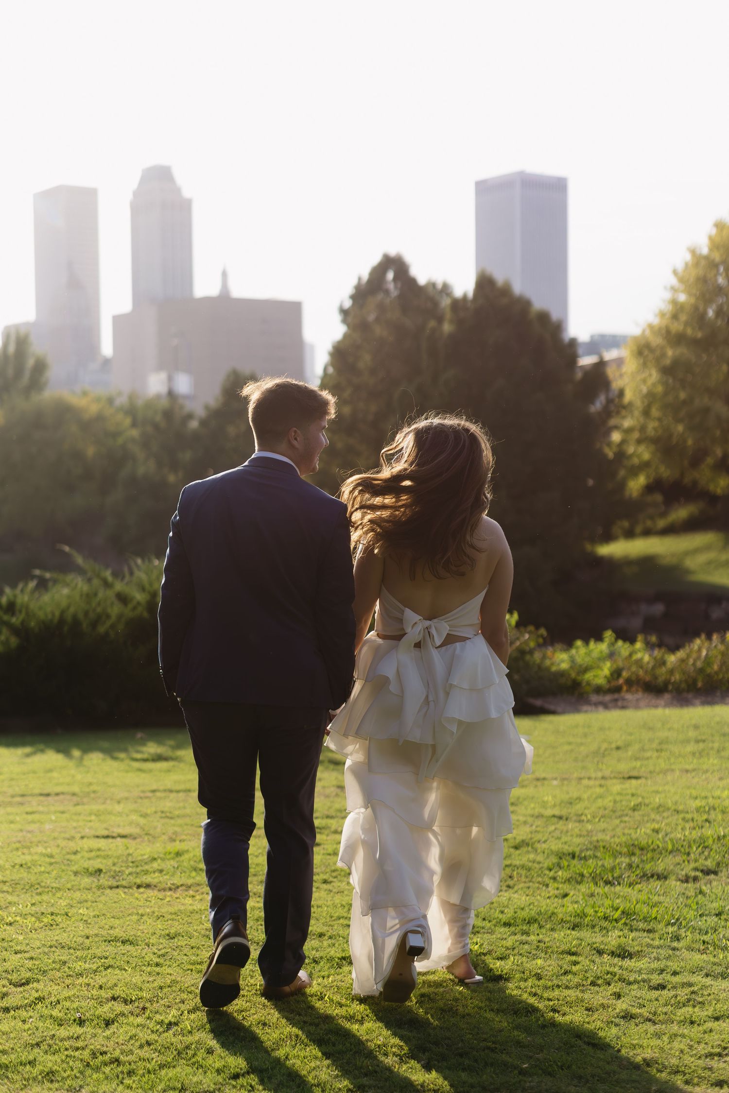 Newlyweds walk hand in hand towards a city skyline at sunset.