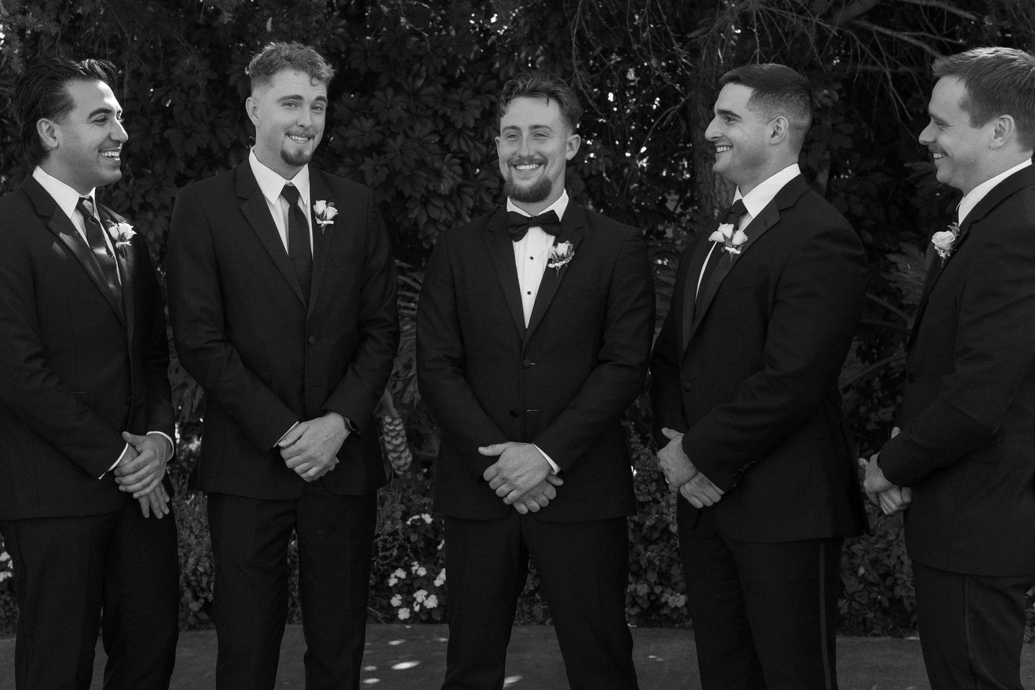 Groomsmen in black tuxedos stand together smiling at an outdoor wedding.