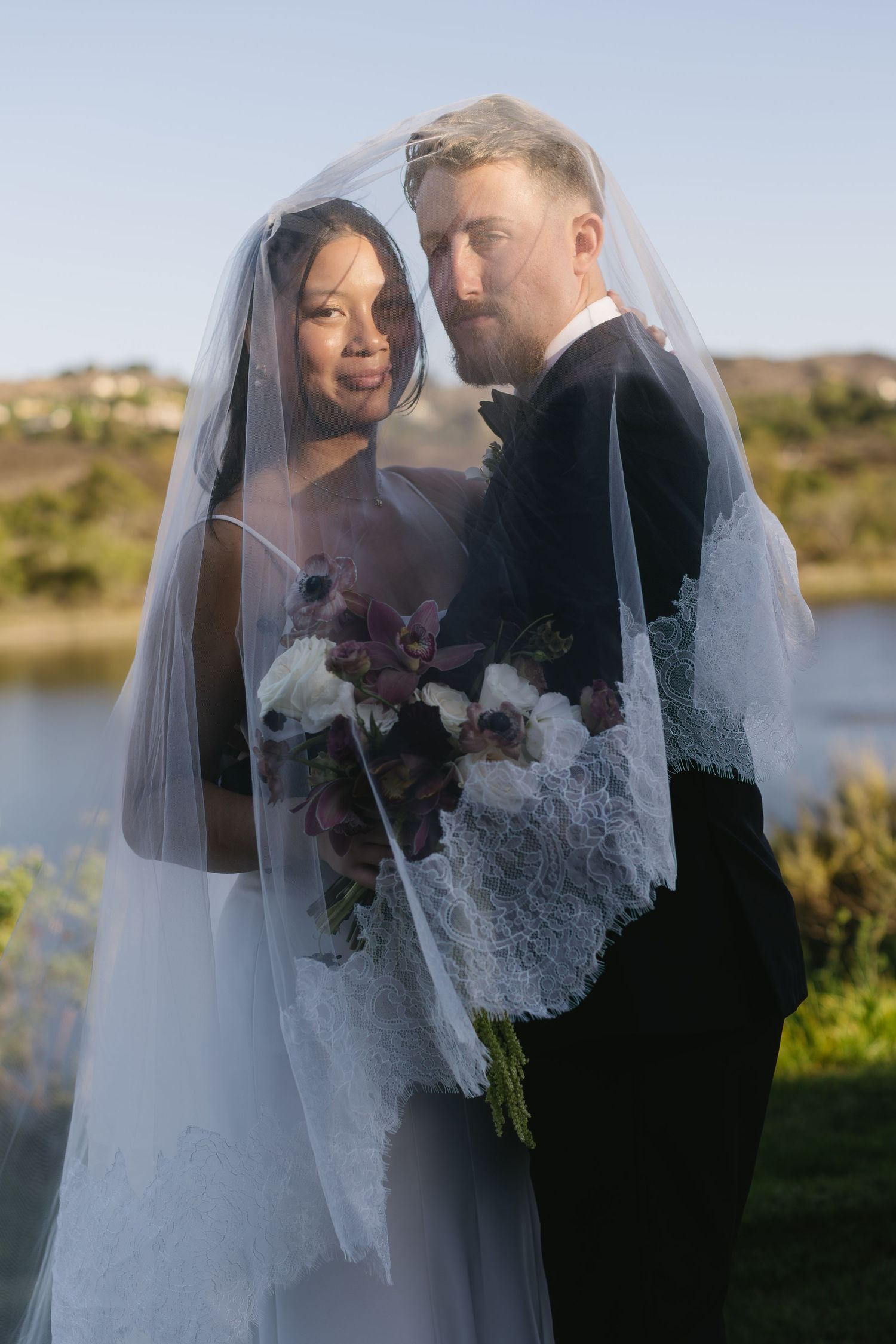 Romantic wedding portrait with couple embracing under a flowing veil against a lakeside backdrop.