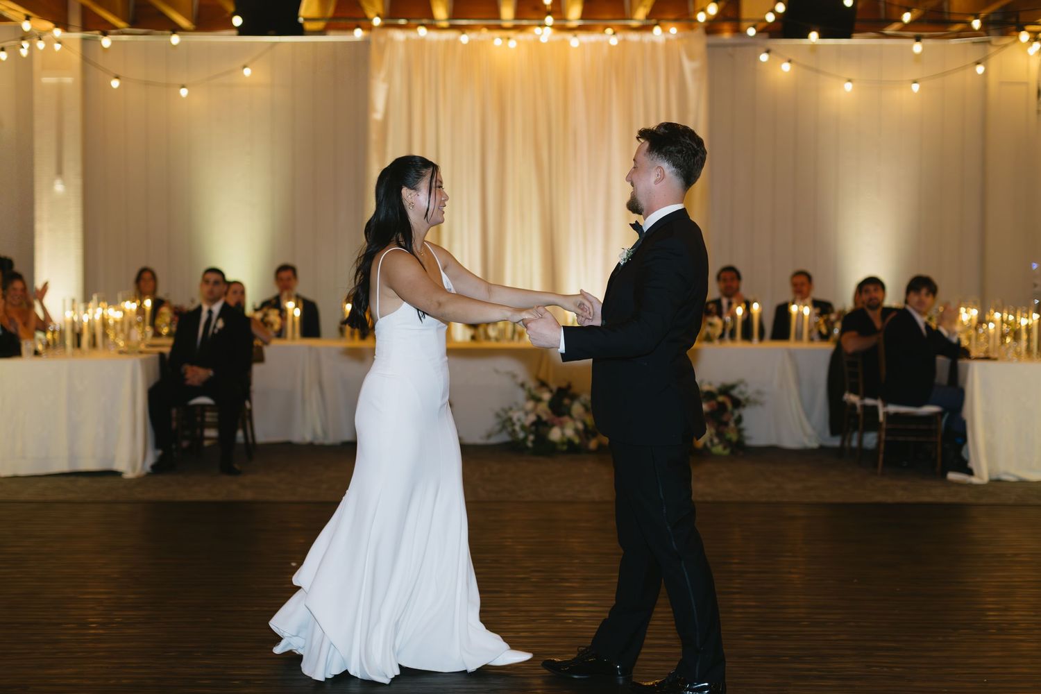 Wedding couple's first dance in an elegant ballroom with string lights and white decor.