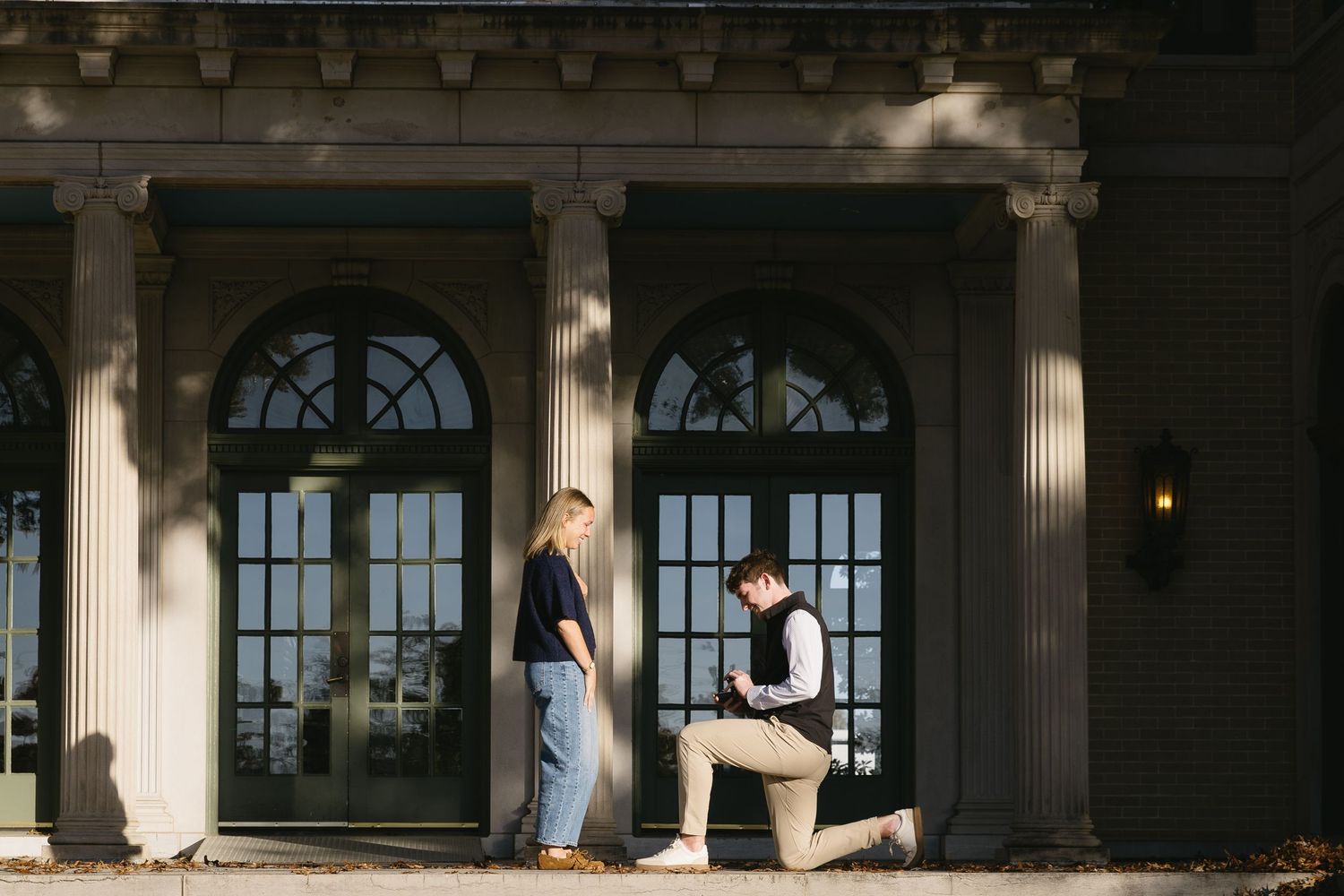A person kneels down to propose in an elegant hallway with arched windows and columns.