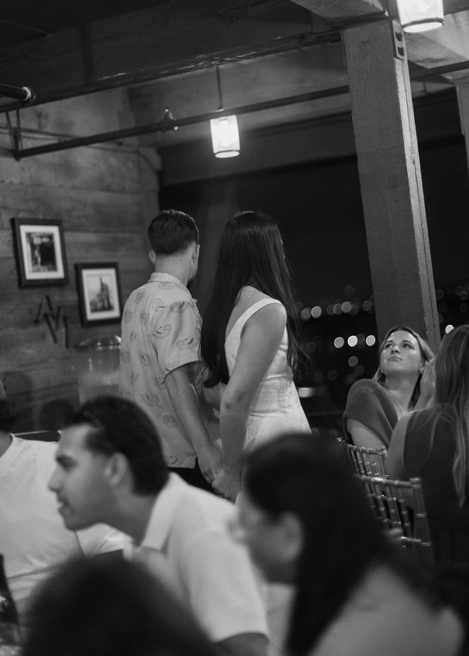 Wedding reception guests mingle and celebrate in a rustic indoor venue with wooden beams.