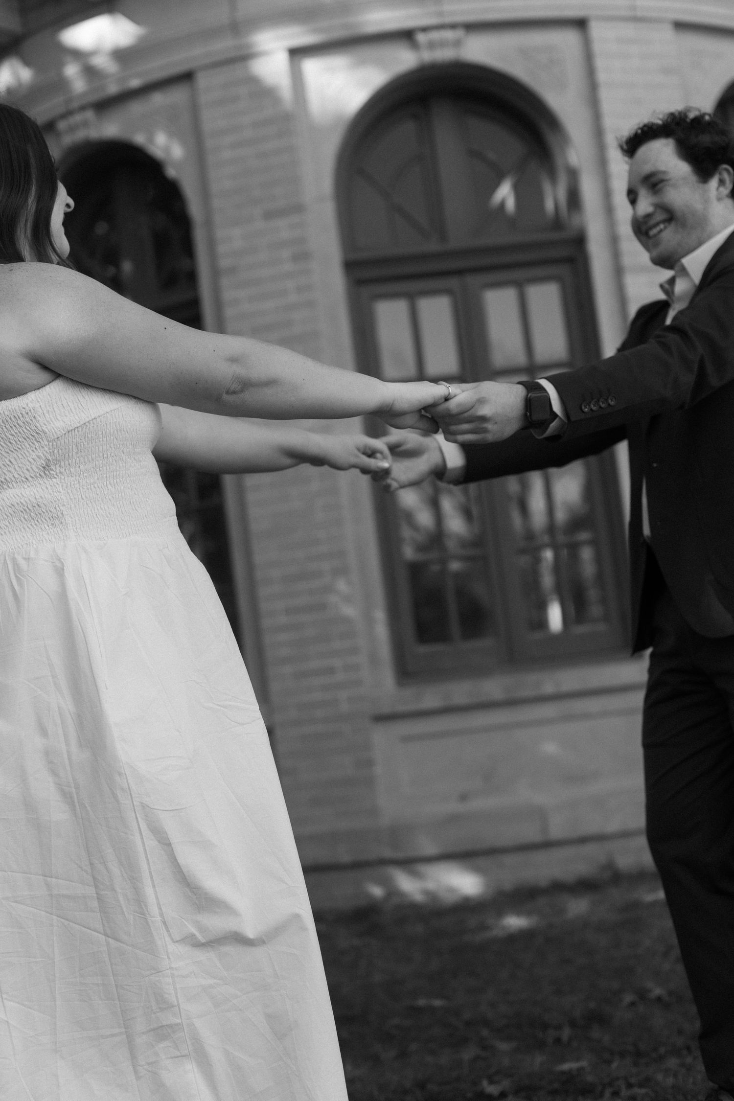 A black and white photo of a couple dancing and holding hands in front of a brick building.