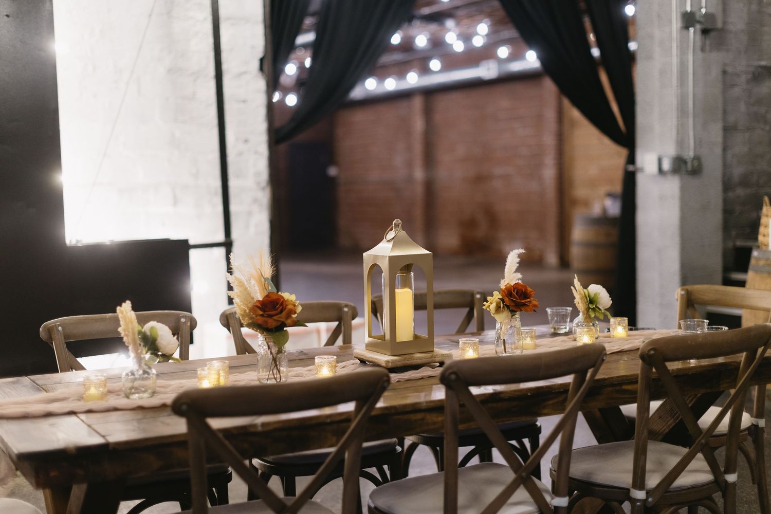 Elegant farmhouse table setting with lanterns and cross-back chairs in a rustic venue.