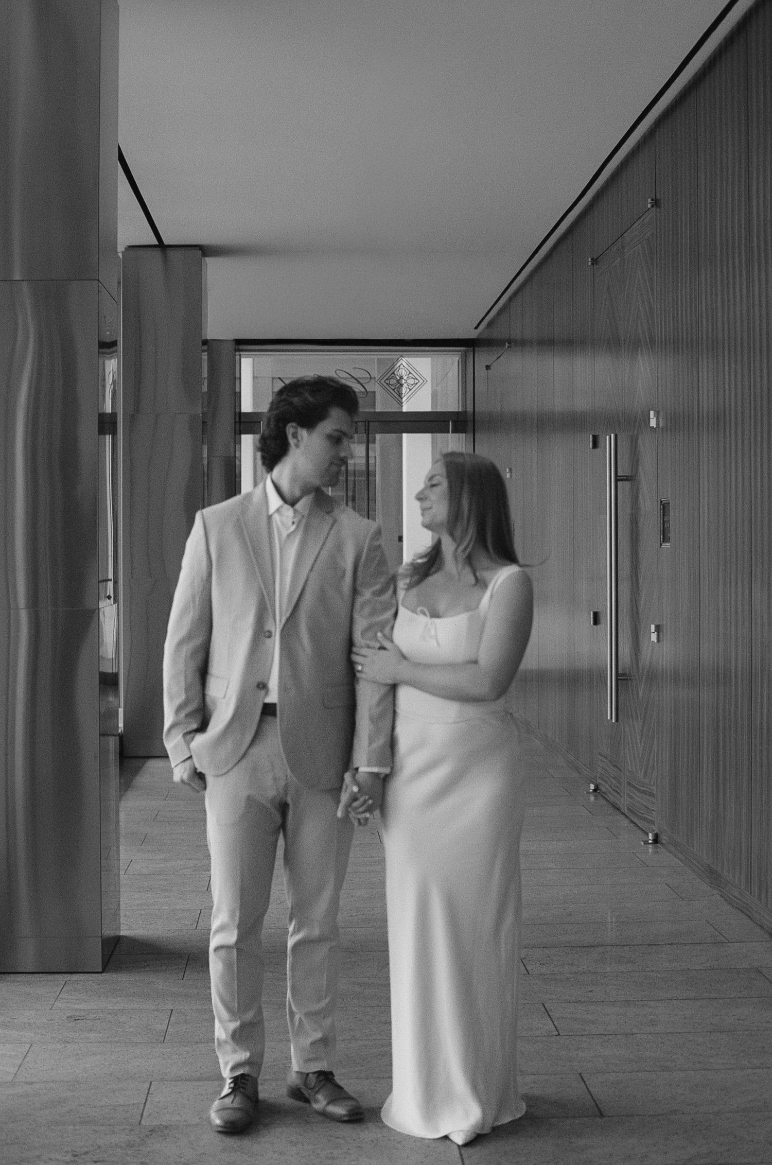 A black and white photo of a couple in formal wear standing together in a hallway.
