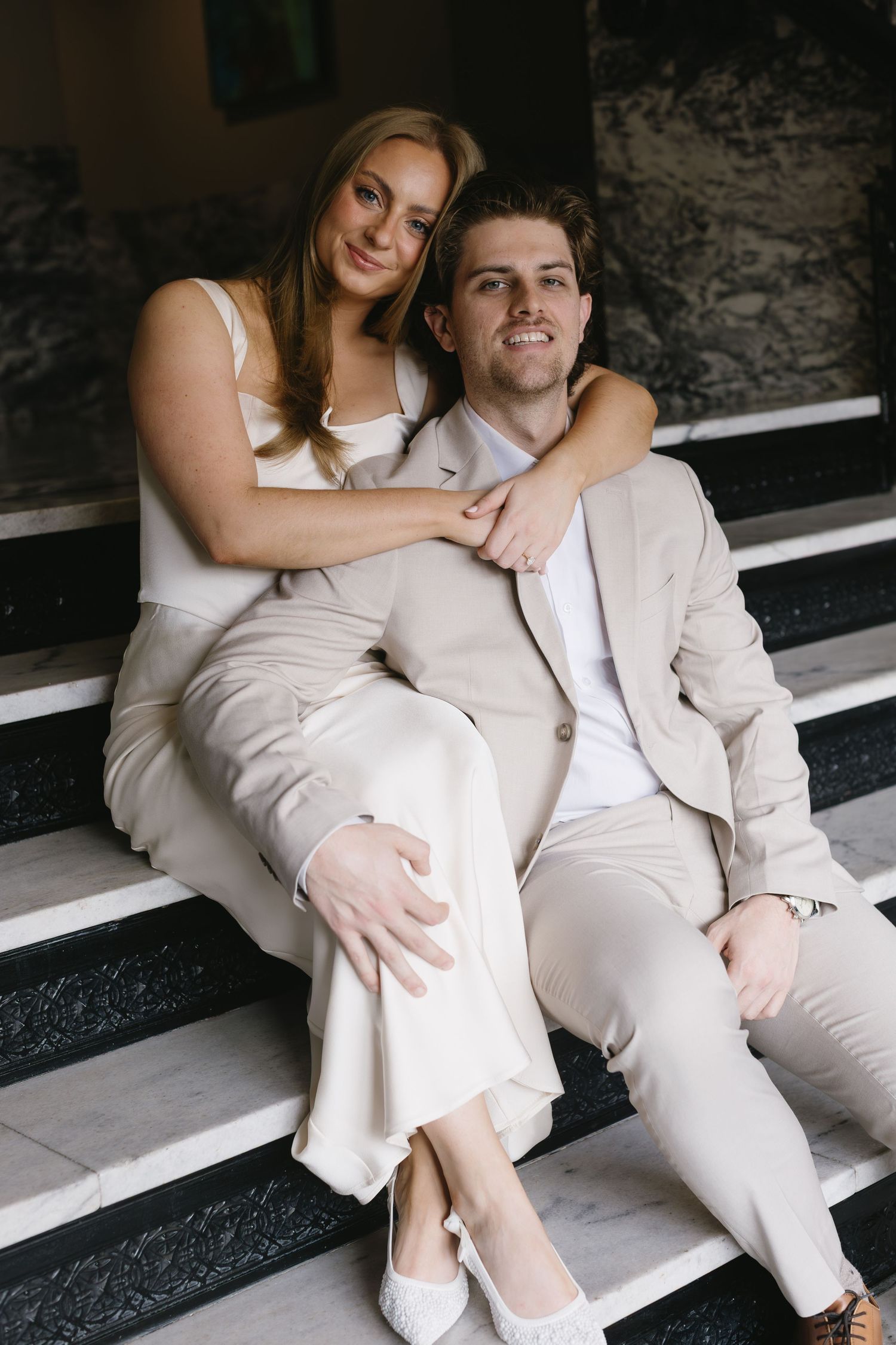 A couple in matching white and beige outfits sits intimately on stone steps.