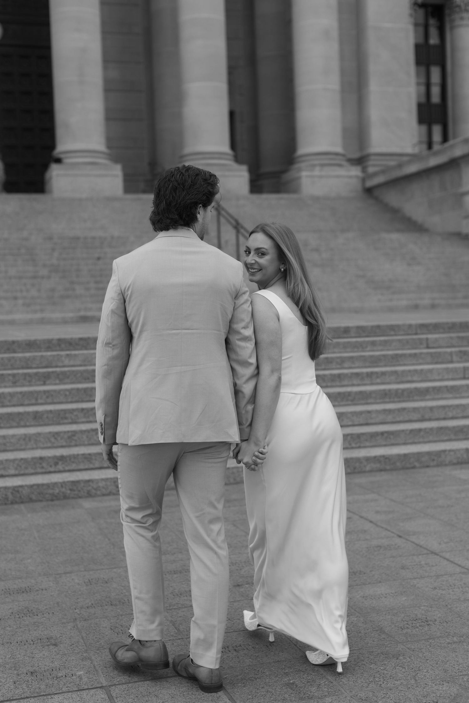 A couple in formal attire walks together along stone steps of a grand building.