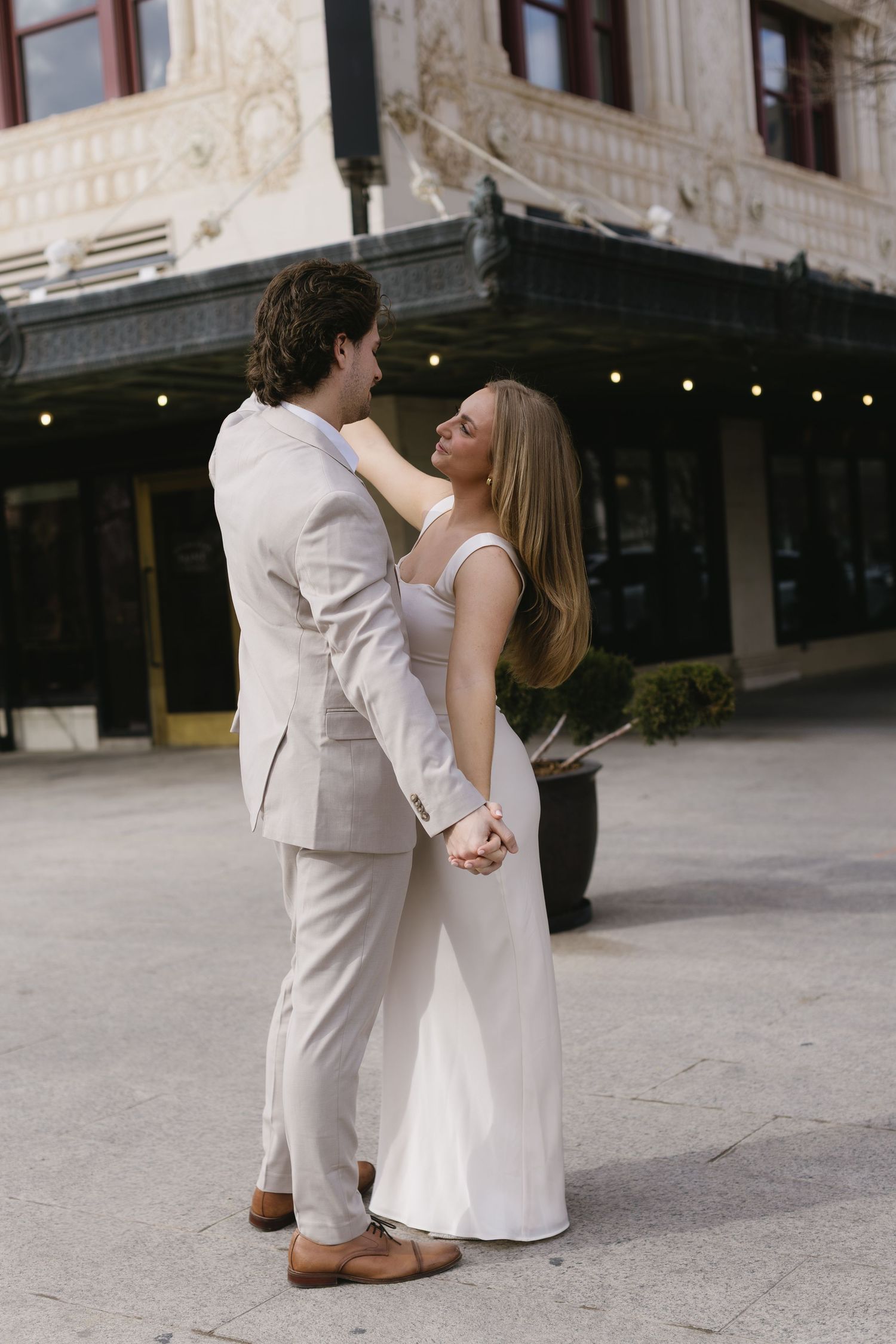 Romantic couple in light-colored formal attire dancing on urban plaza.