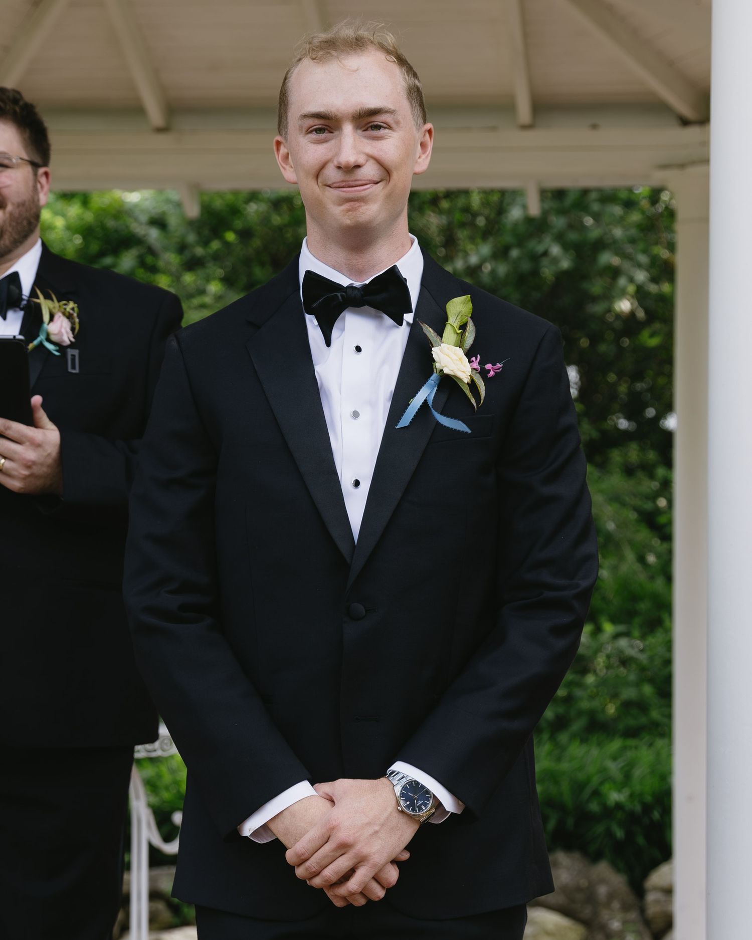 A formal wedding party member in a black tuxedo with boutonniere at an outdoor venue.