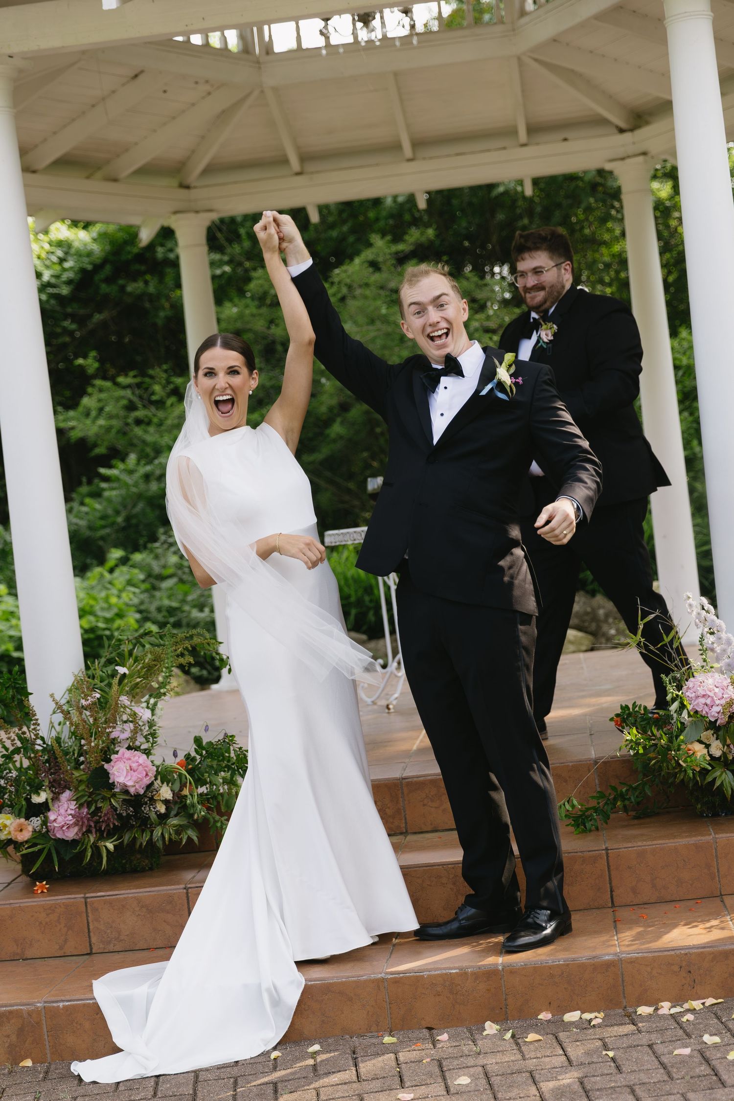 Wedding party celebrates joyfully under a white gazebo surrounded by greenery and flowers during outdoor ceremony.
