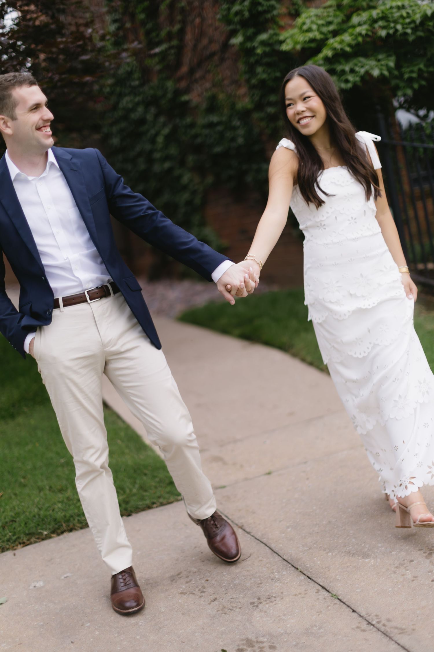 A joyful couple in wedding attire hold hands while walking along a pathway surrounded by greenery.