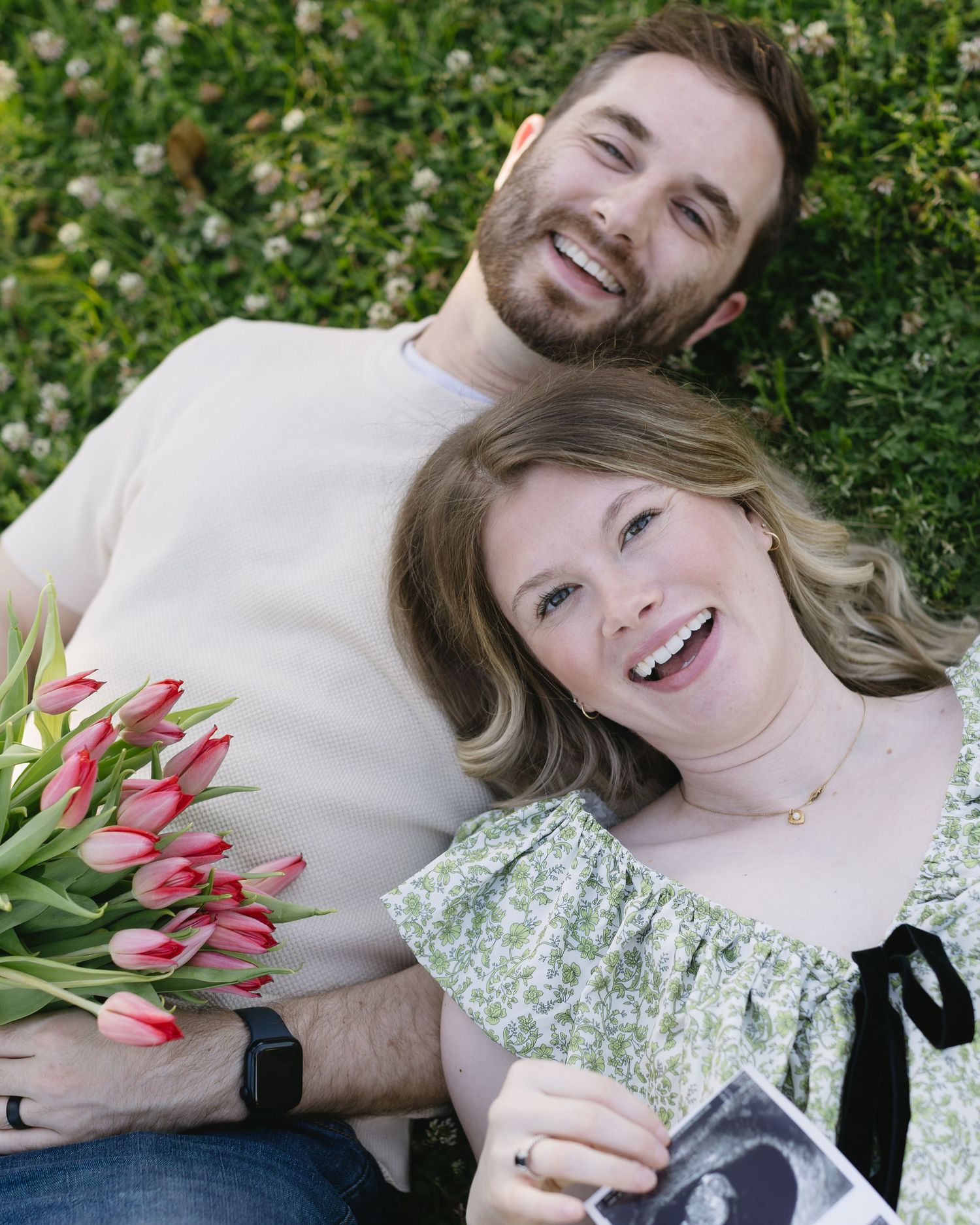 A joyful outdoor maternity photoshoot with tulip bouquet and sonogram photo against green foliage.
