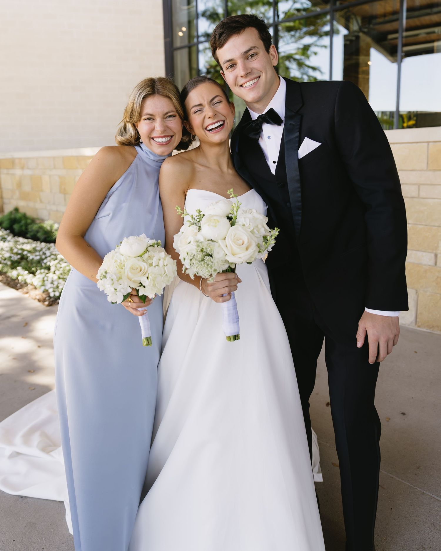 Wedding party poses together in formal attire against stone wall and window backdrop.