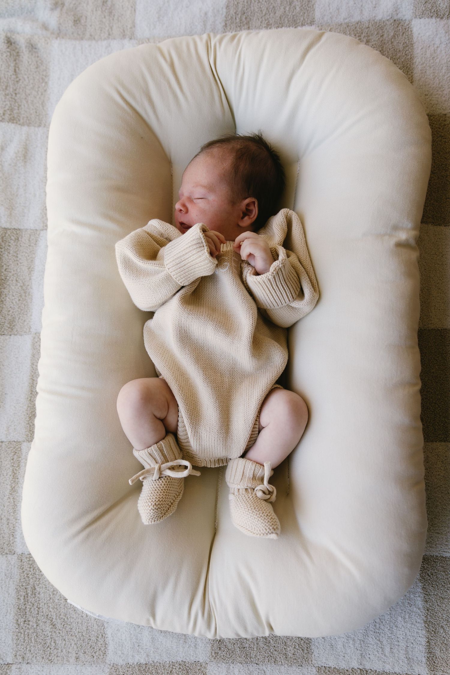 A peaceful newborn in cream-colored clothing rests on a white cushioned lounger.