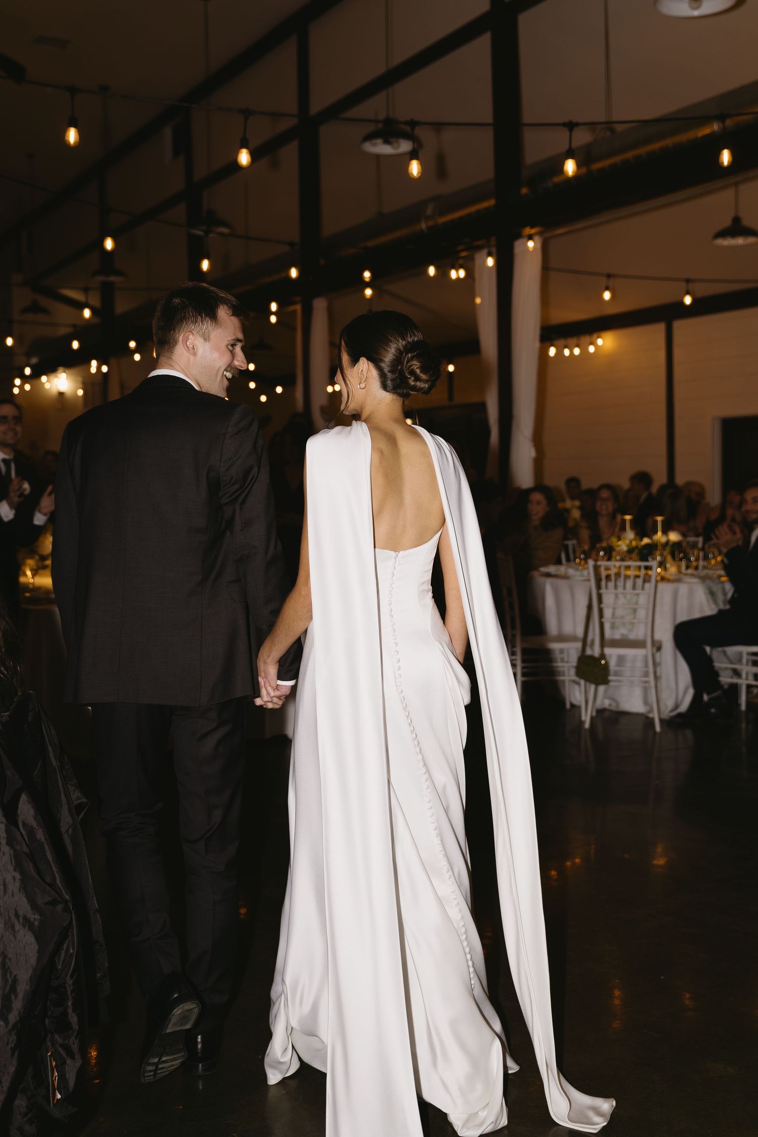 A couple in formal attire stands in a reception venue with ambient string lighting overhead.