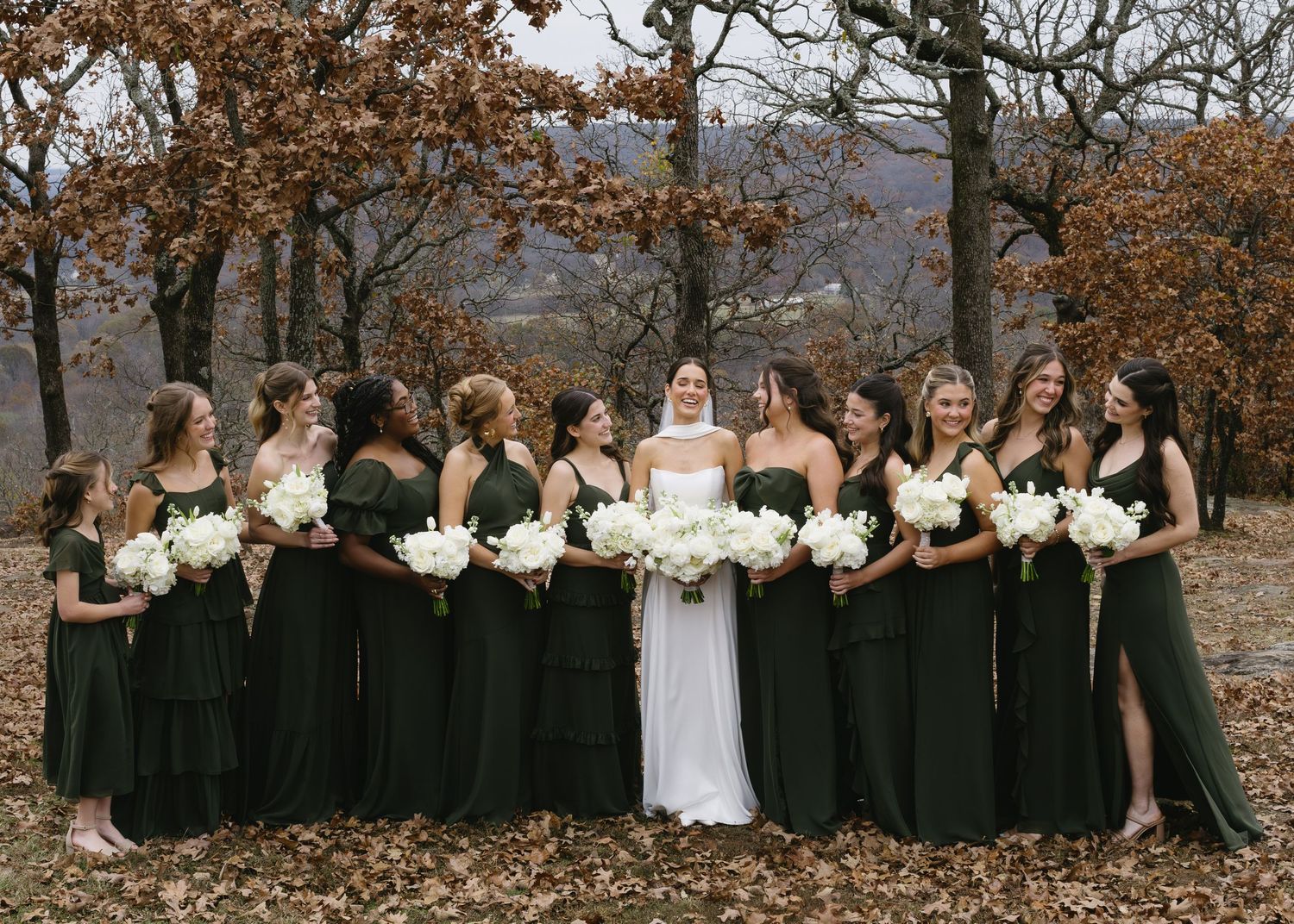Wedding party in black dresses poses with bride against autumn foliage backdrop.